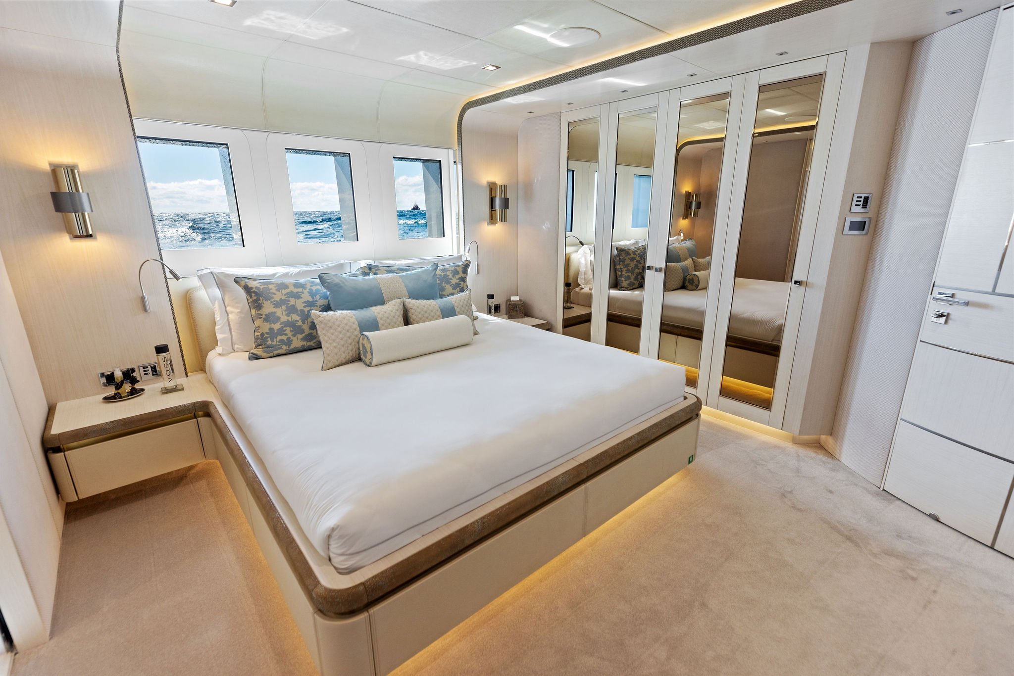 Luxurious bedroom with a view of the ocean through windows, white bed with decorative pillows, mirrored closet doors, and modern lighting.