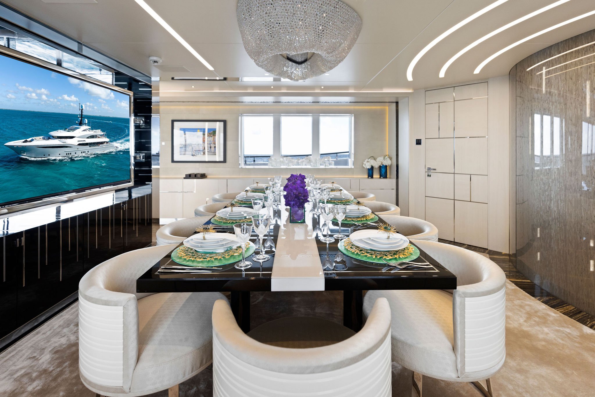 A luxurious yacht dining room with a black dining table set for eight, surrounded by cream-colored curved chairs. The table has white plates, gold flatware, and wine glasses, with a purple floral centerpiece. A large window at the back shows a boat o