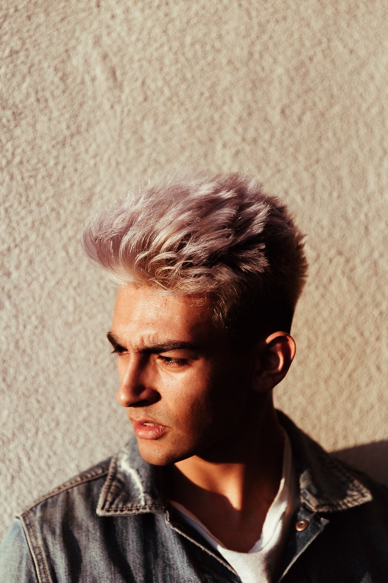 Young man with platinum blonde hair and denim jacket looking down against a textured beige wall.