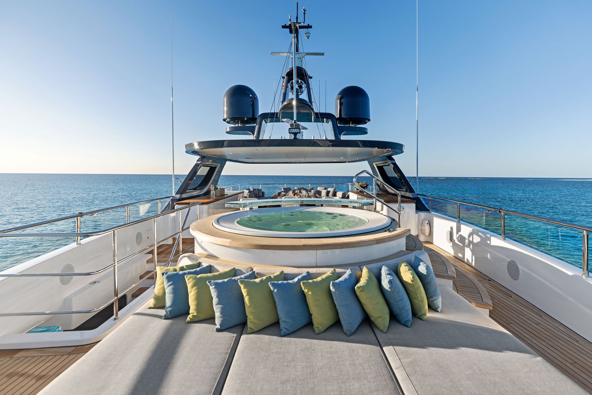 On the deck of a luxury yacht, overlooking a calm ocean, there is a hot tub surrounded by colorful cushions, with seating and modern technology visible on the yacht's upper deck.