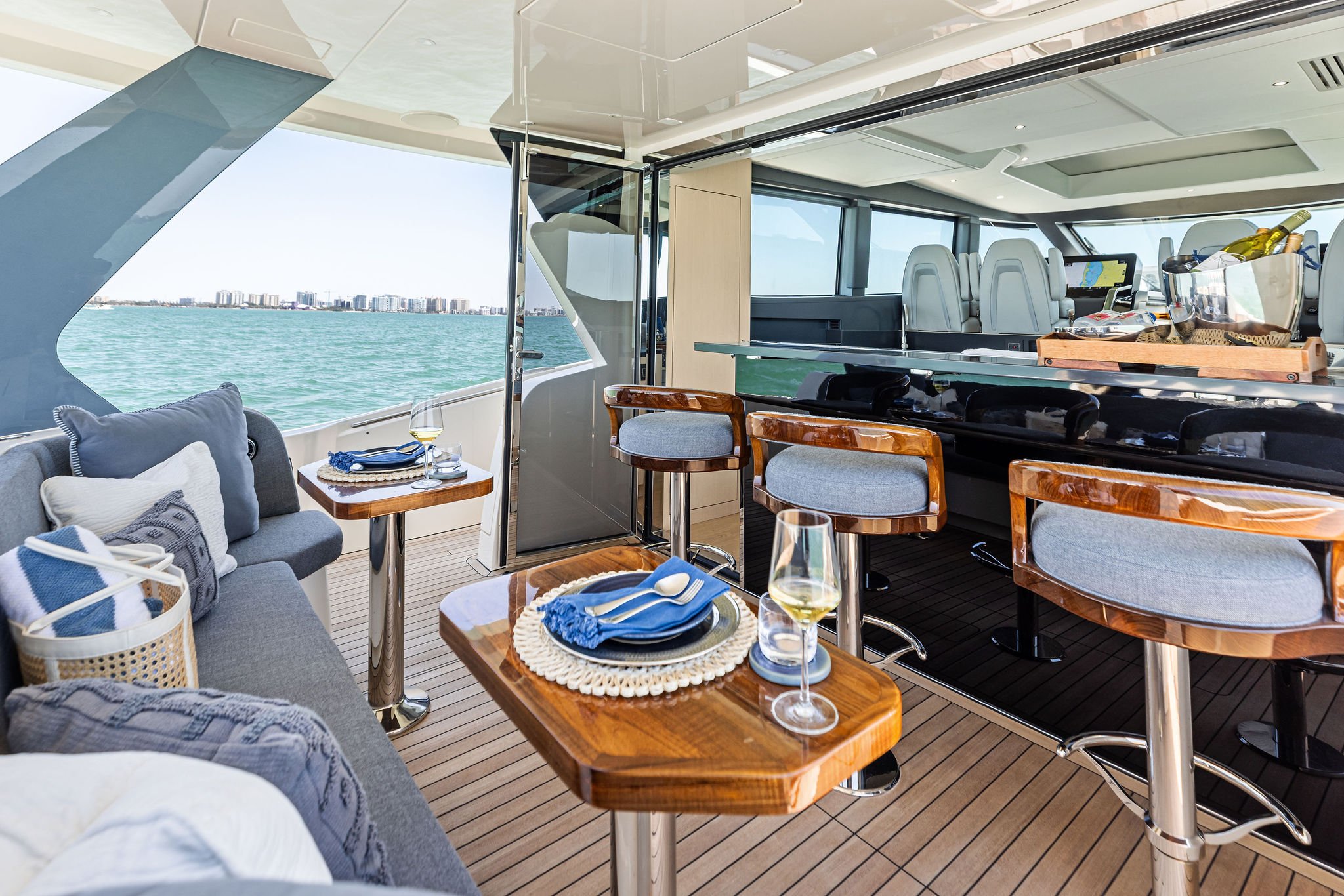 The interior of a yacht with a seating area, tables set with plates, utensils, and wine glasses, a bar area with bottles of wine and glasses, and large windows showing water and city skyline in the background.