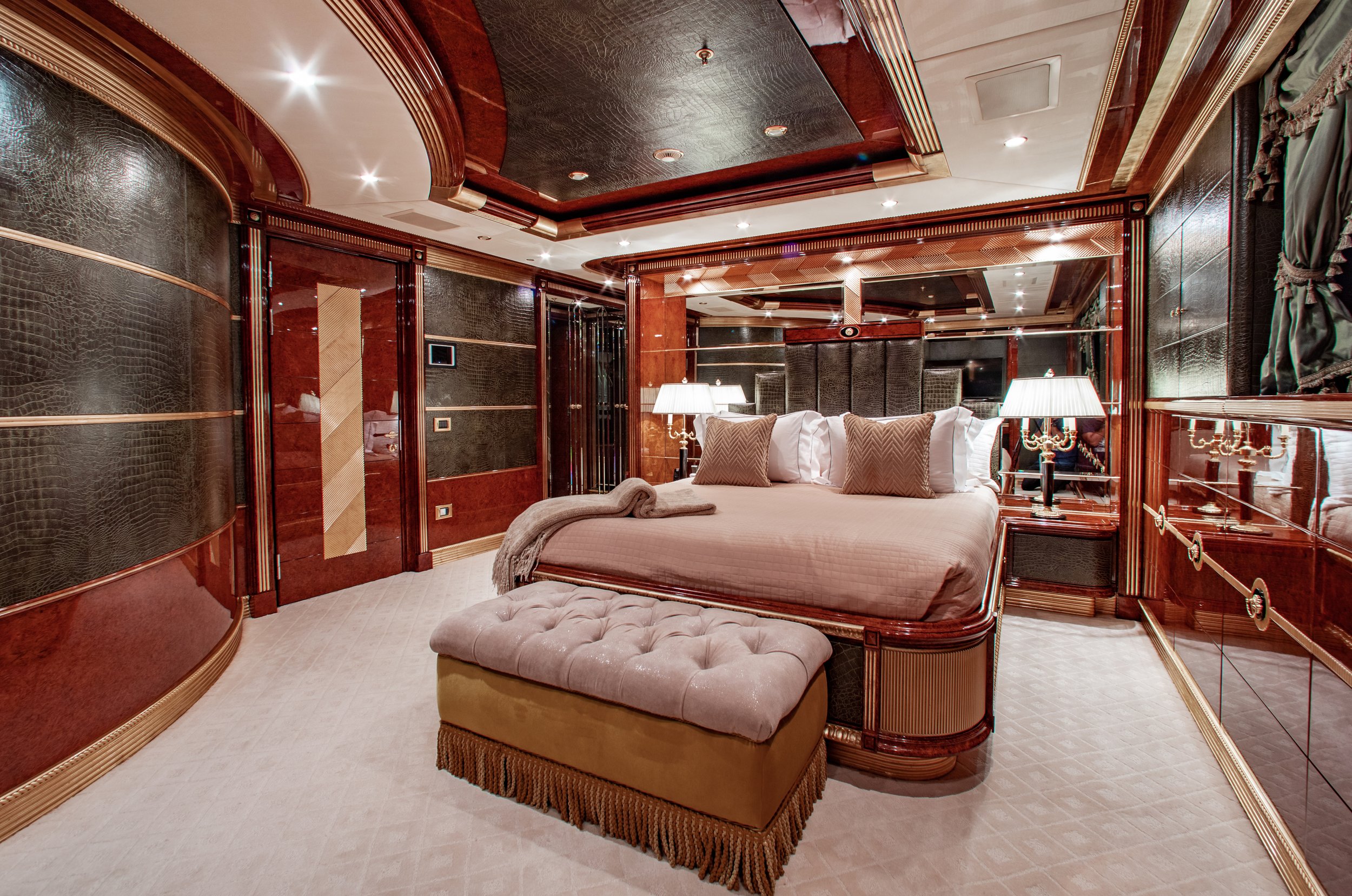 Luxurious hotel bedroom with a large bed, upholstered bench, mirrored walls, dark wood paneling, and elegant lamps.