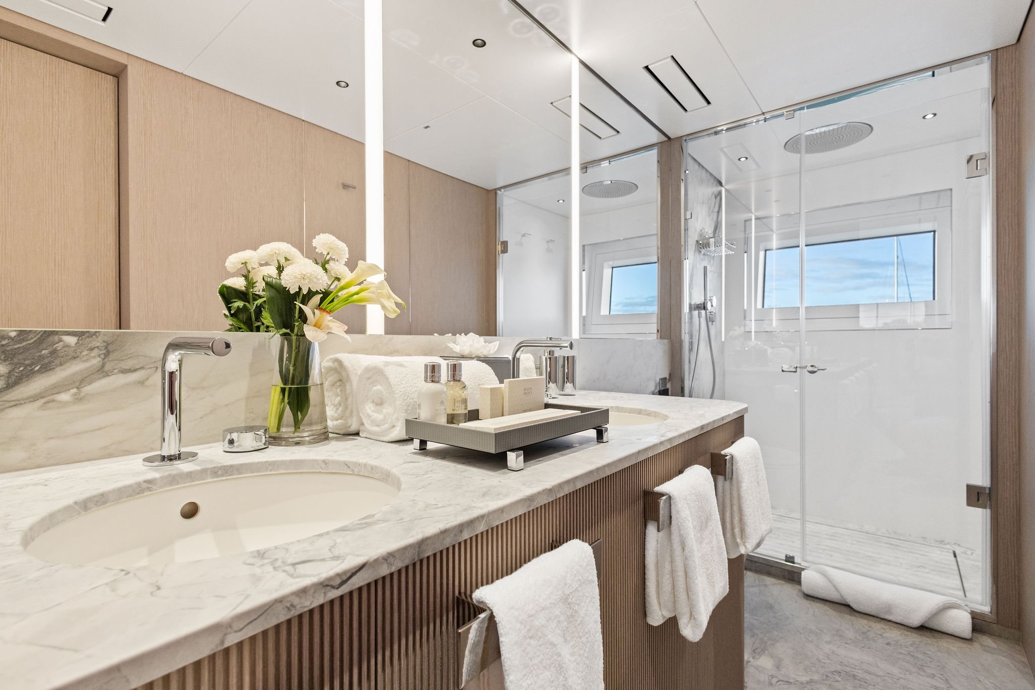 Modern bathroom with marble countertop, a vase of white flowers, rolled towels, toiletries, a large mirror, and a glass-enclosed shower with a window showing a blue sky.