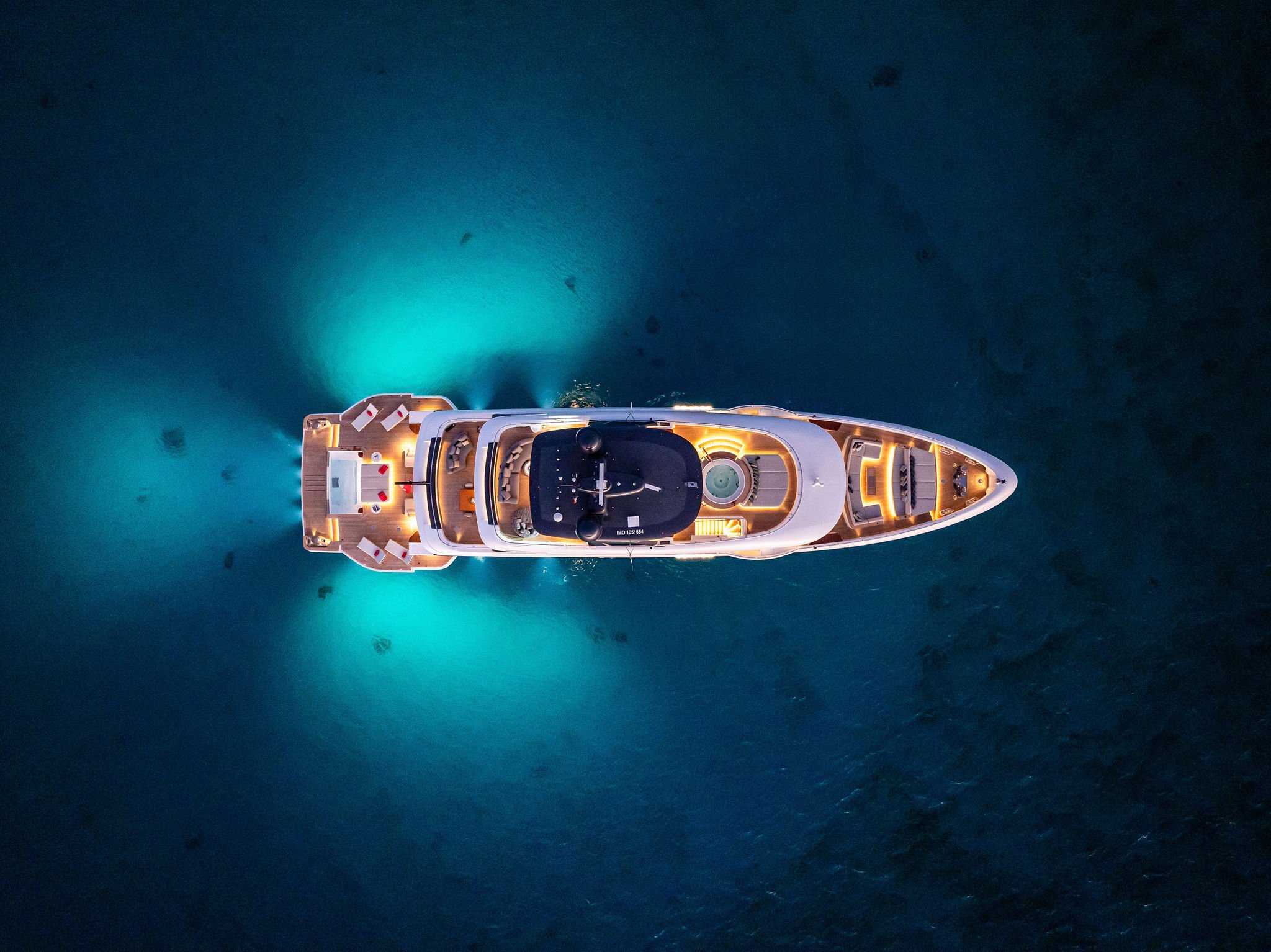 Top-down aerial view of a luxury yacht illuminated at night, floating in calm deep blue water.