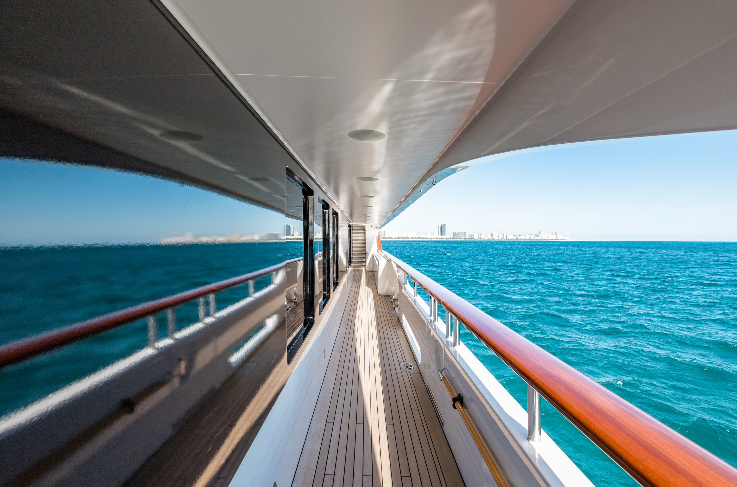 View from the deck of a yacht showing wooden flooring, glass windows, and a distant city skyline over blue ocean water.