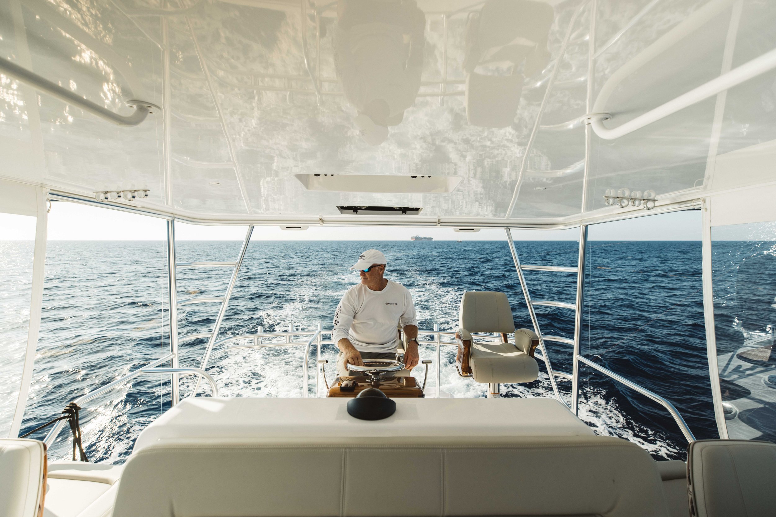 A man wearing a white cap, sunglasses, and a white long-sleeve shirt is steering a boat on the open ocean. The boat has white seats and a white ceiling, with a view of the blue ocean and distant ships on the horizon.