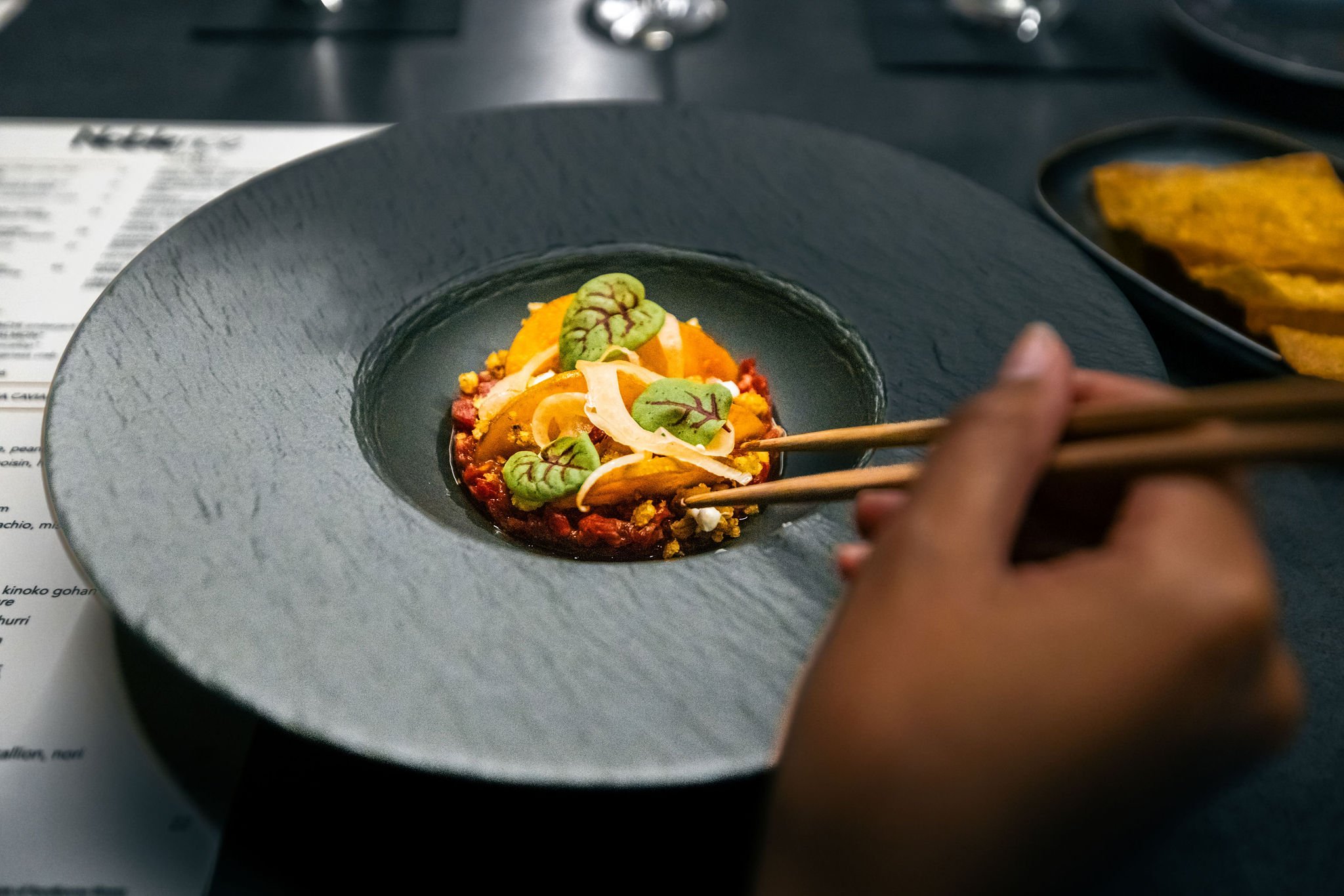 Person using chopsticks to pick up food from a black textured plate containing an Asian-style dish with vegetables, herbs, and sauce, with a small black bowl of crispy chips or crackers in the background.