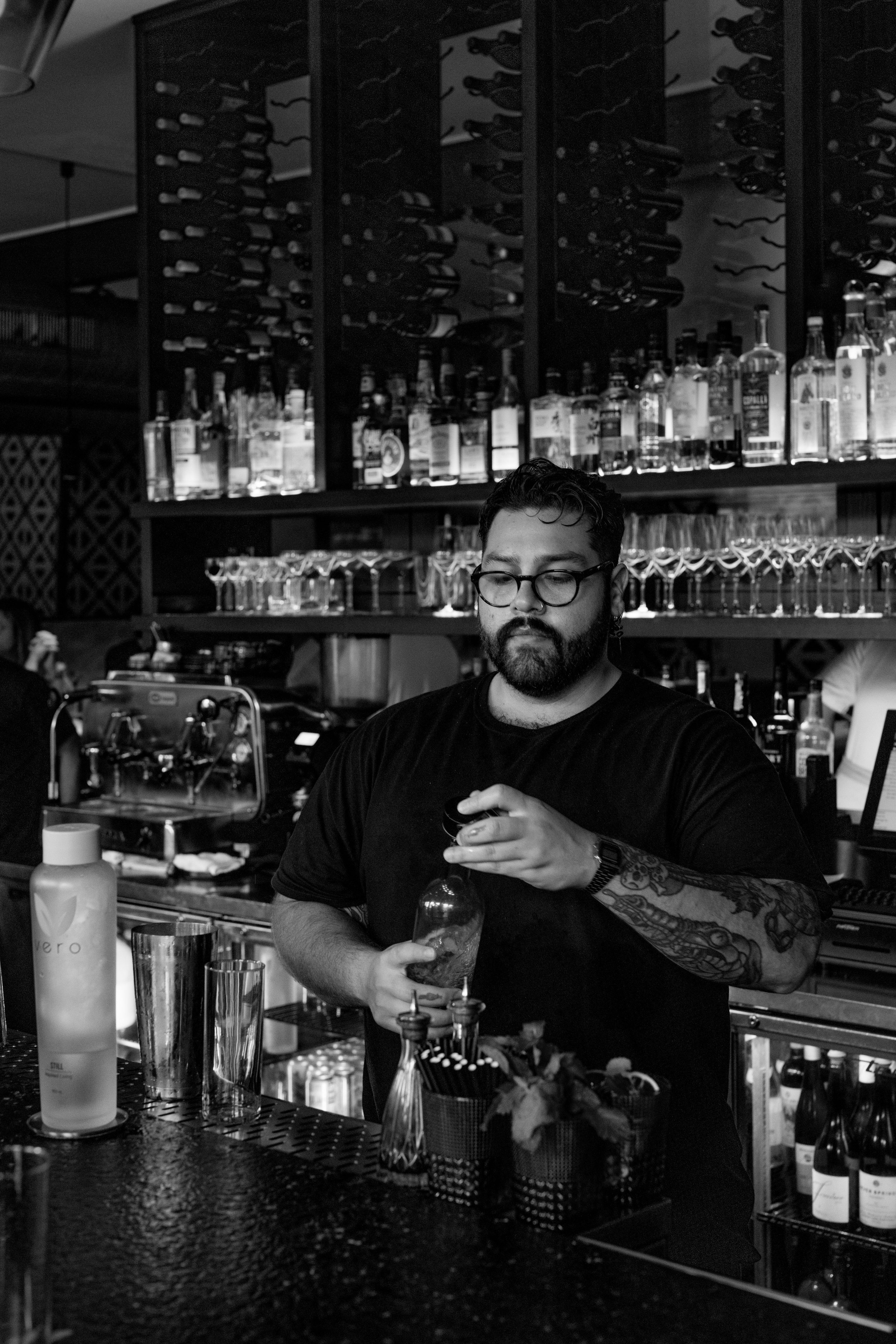 A bartender with glasses and tattoos pouring a drink at a bar with bottles of alcohol and bar tools behind him, in black and white.
