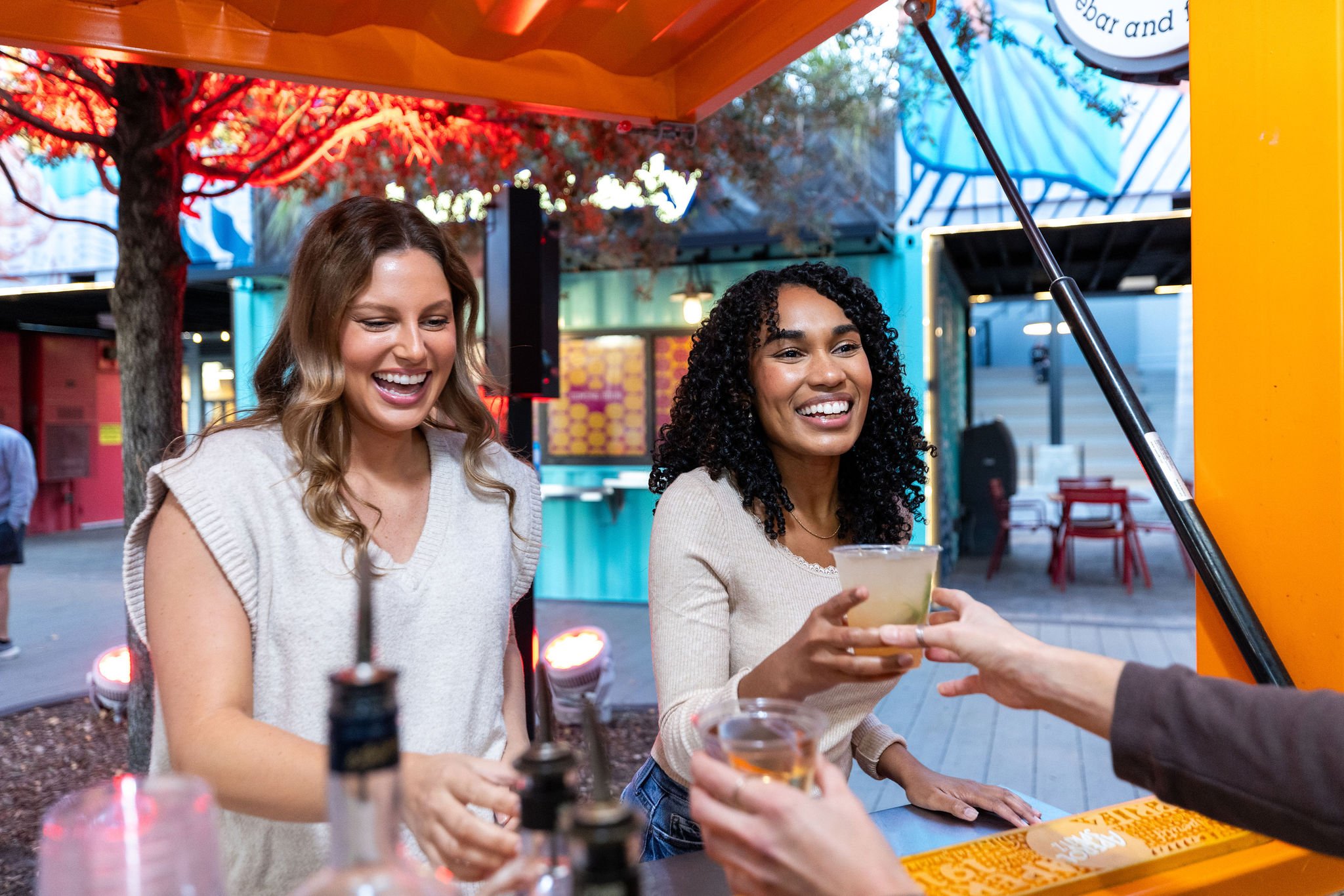 Two women joyfully receive and enjoy drinks from a bar at an outdoor food market during the evening, with colorful background and orange and blue decor.