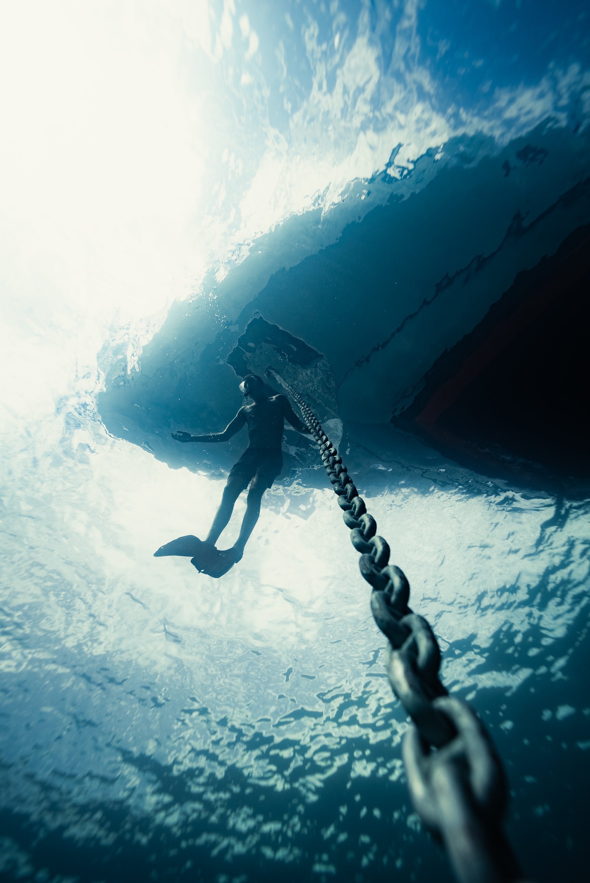 A person with fins and a mask attached to a chain, swimming underwater near a large rock formation or boat, with clouds visible through the water's surface.