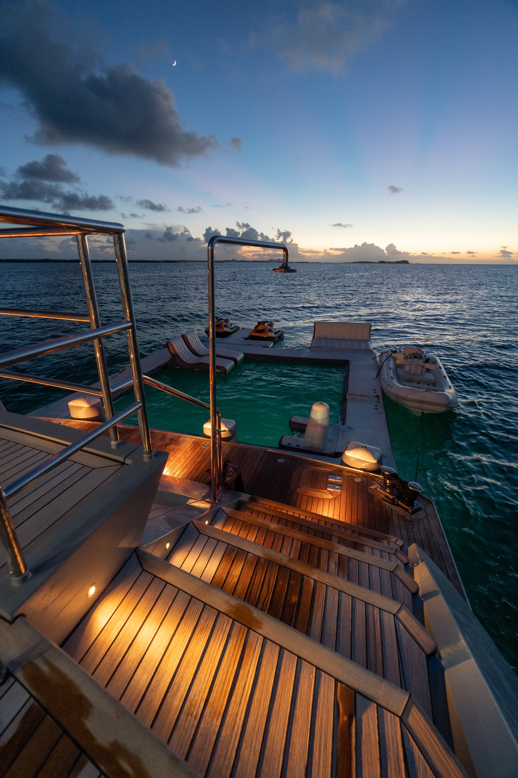 Luxury yacht deck with wooden sun loungers and stairs leading down to a hot tub, illuminated by small lights, at dusk with a calm ocean and cloudy sky in the background.