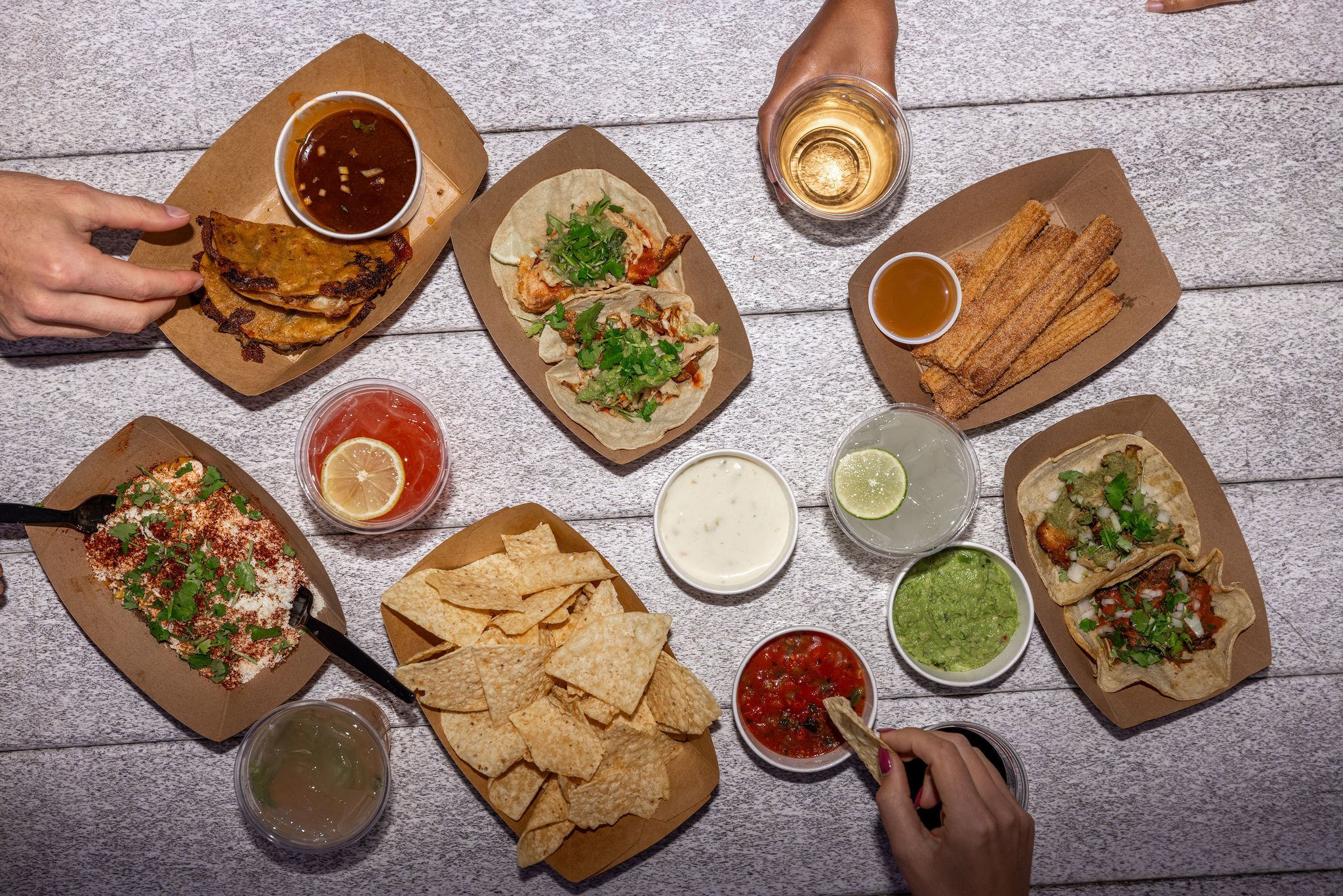 An overhead view of various Mexican dishes and drinks on a wooden surface, including tacos, churros, chips with salsa, queso, guacamole, and drinks like beer, tequila, and water, with hands serving and reaching for the food.
