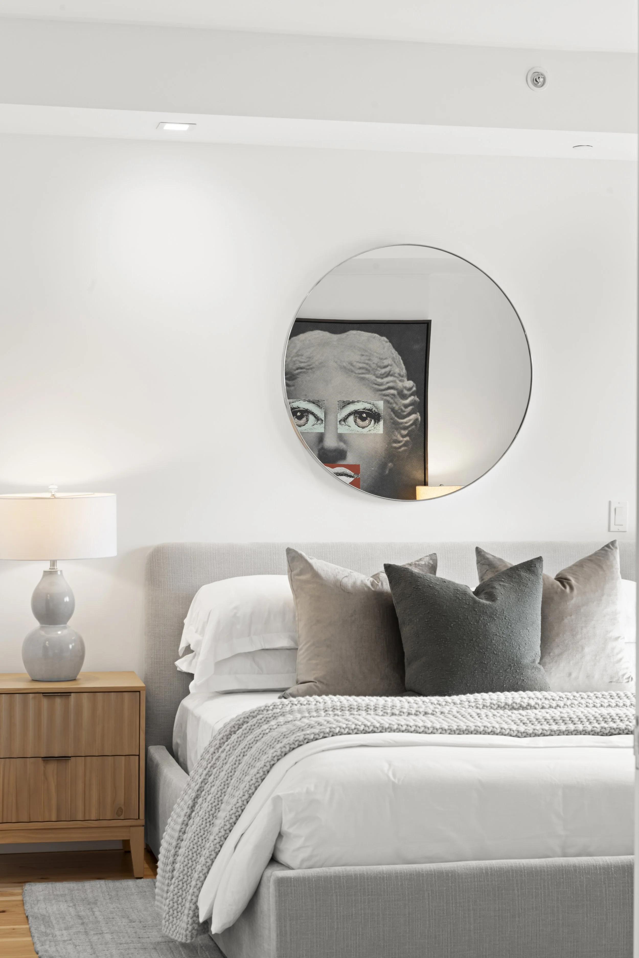 Modern bedroom with a gray upholstered bed, white bedding, and gray throw blanket. A round mirror above the bed reflects artwork. A wooden nightstand holds a ceramic lamp.