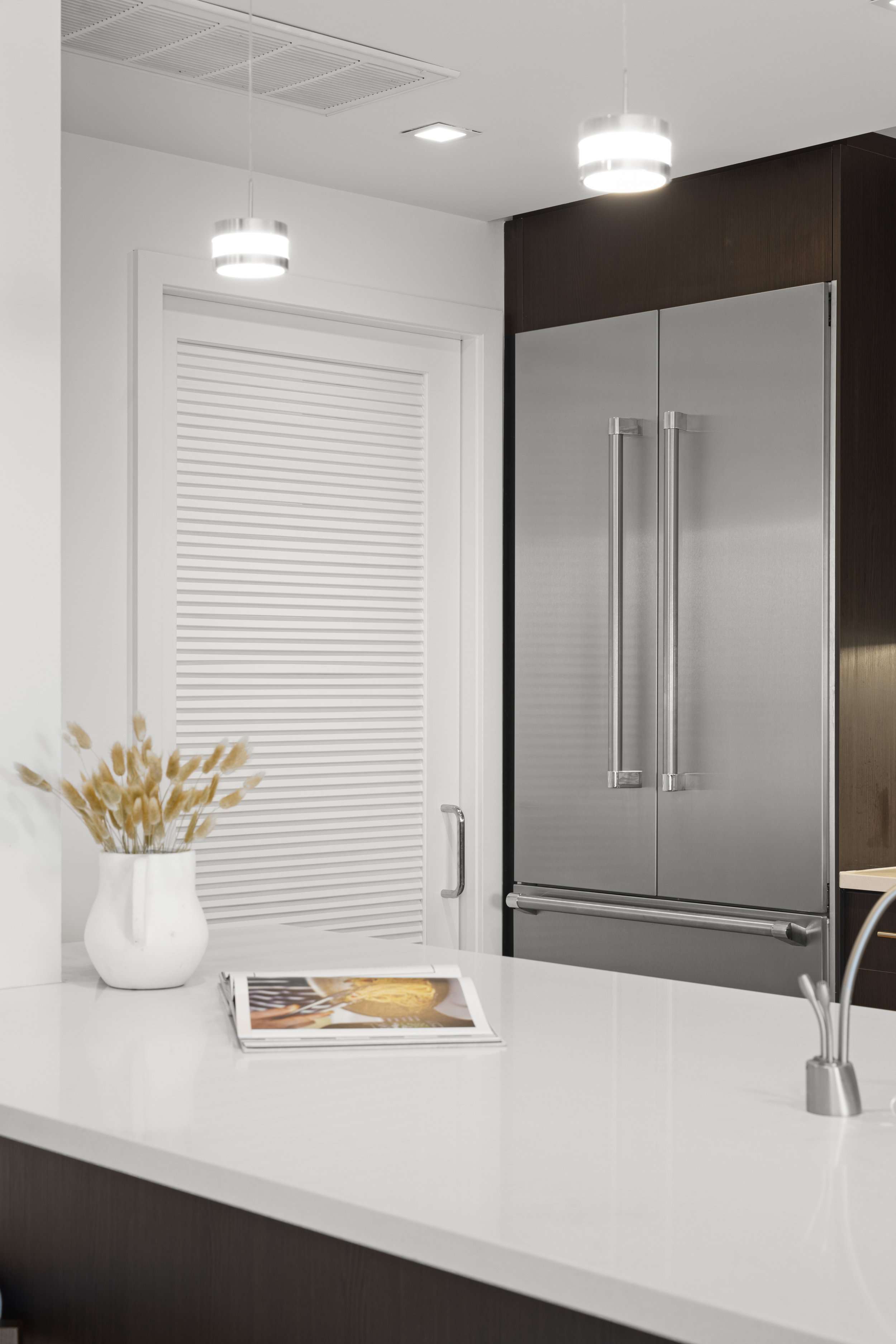 Modern kitchen interior with a stainless steel refrigerator, a white countertop, a vase with dried flowers, and a book on the counter.