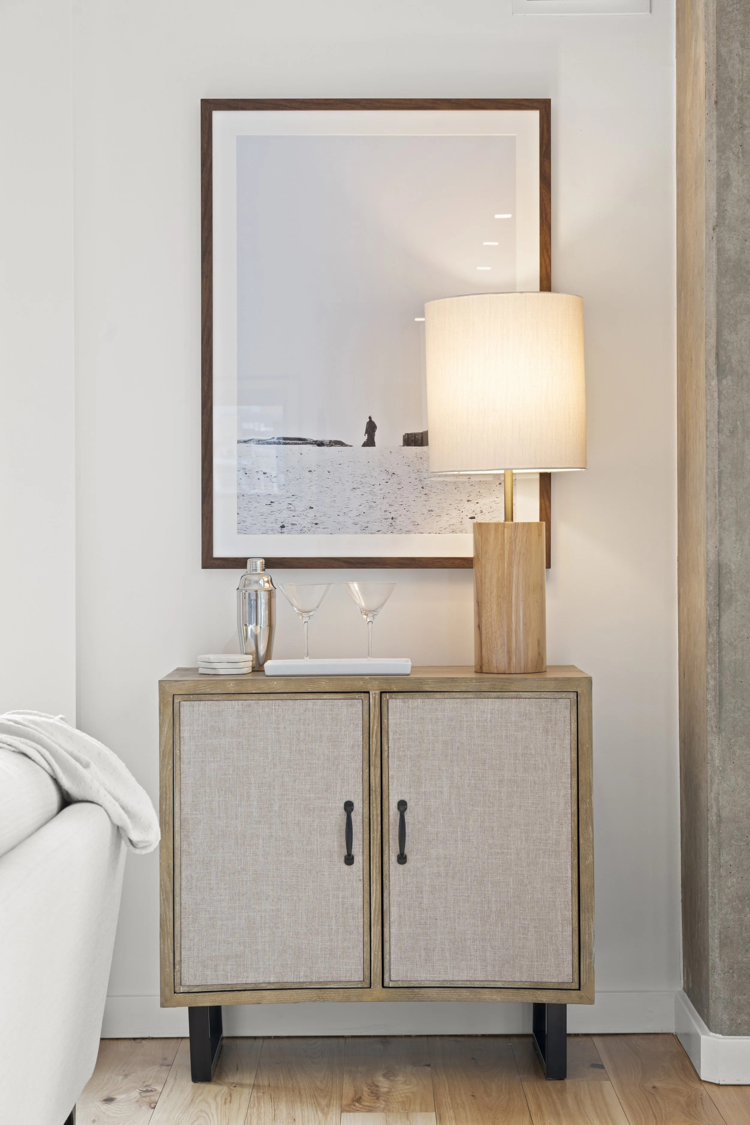 A small cabinet with fabric-covered doors, topped with two martini glasses, a metal cocktail shaker, a framed photograph, and a table lamp, positioned in a modern living room setting.