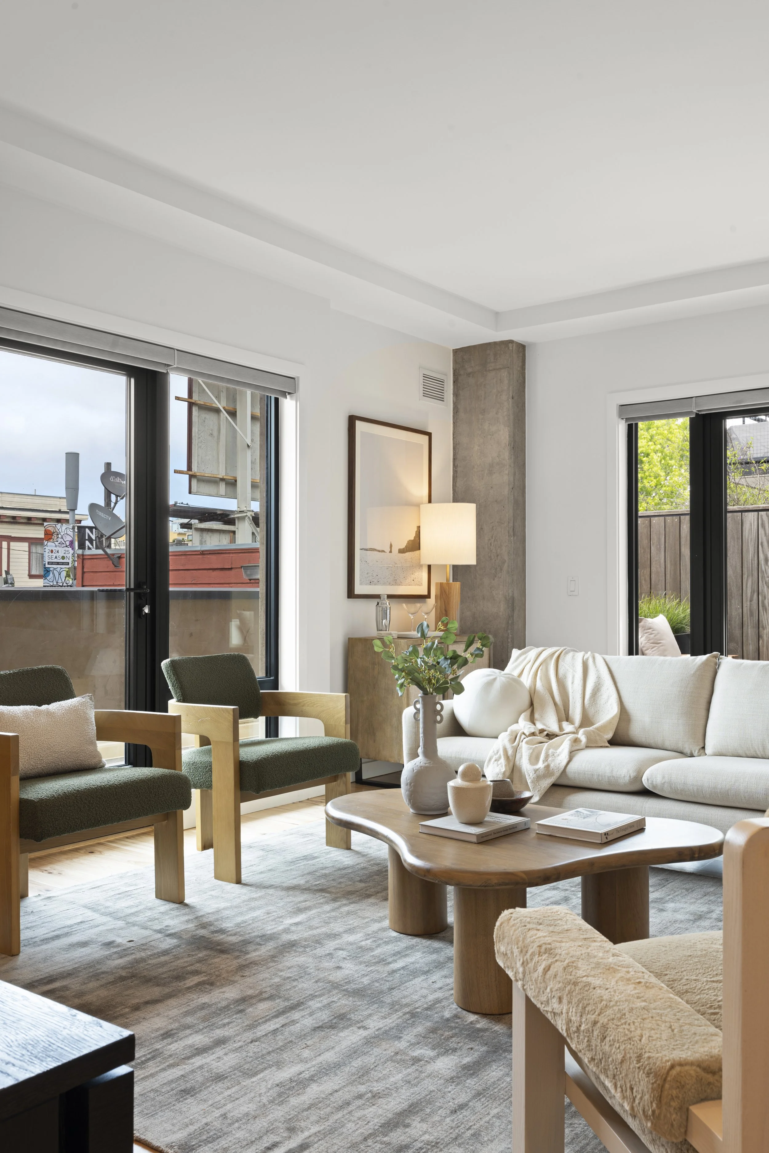Modern living room with large windows, white couch, green and beige chairs, wooden coffee table, and decorative vases. Natural light fills the space, creating a cozy atmosphere.