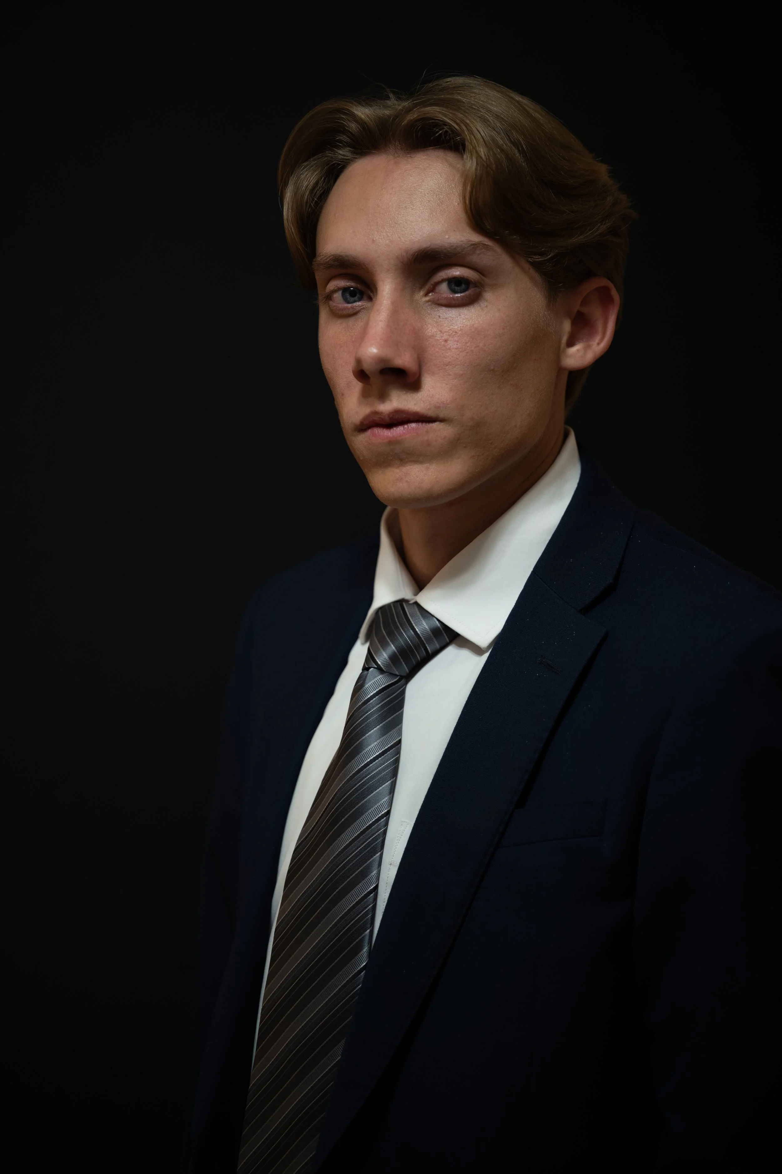 Young man with light brown hair wearing a dark suit, white shirt, and striped tie, standing against a black background.