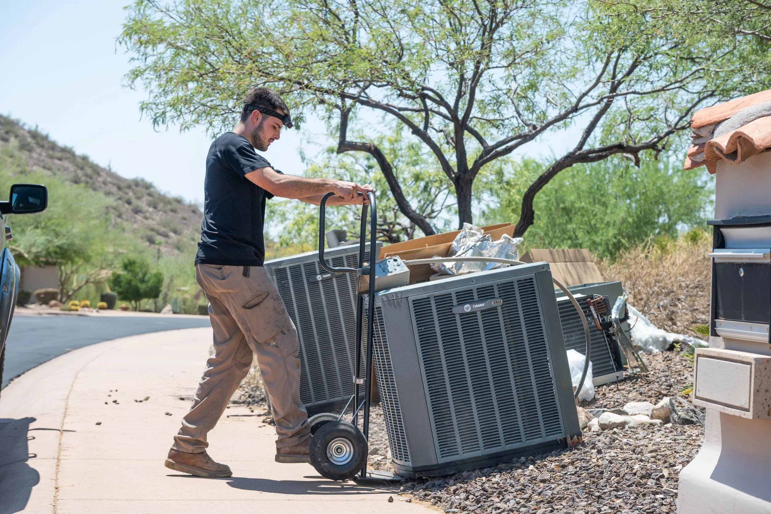 A man pushing a hand truck carrying broken appliance parts next to the curb on a sunny day.