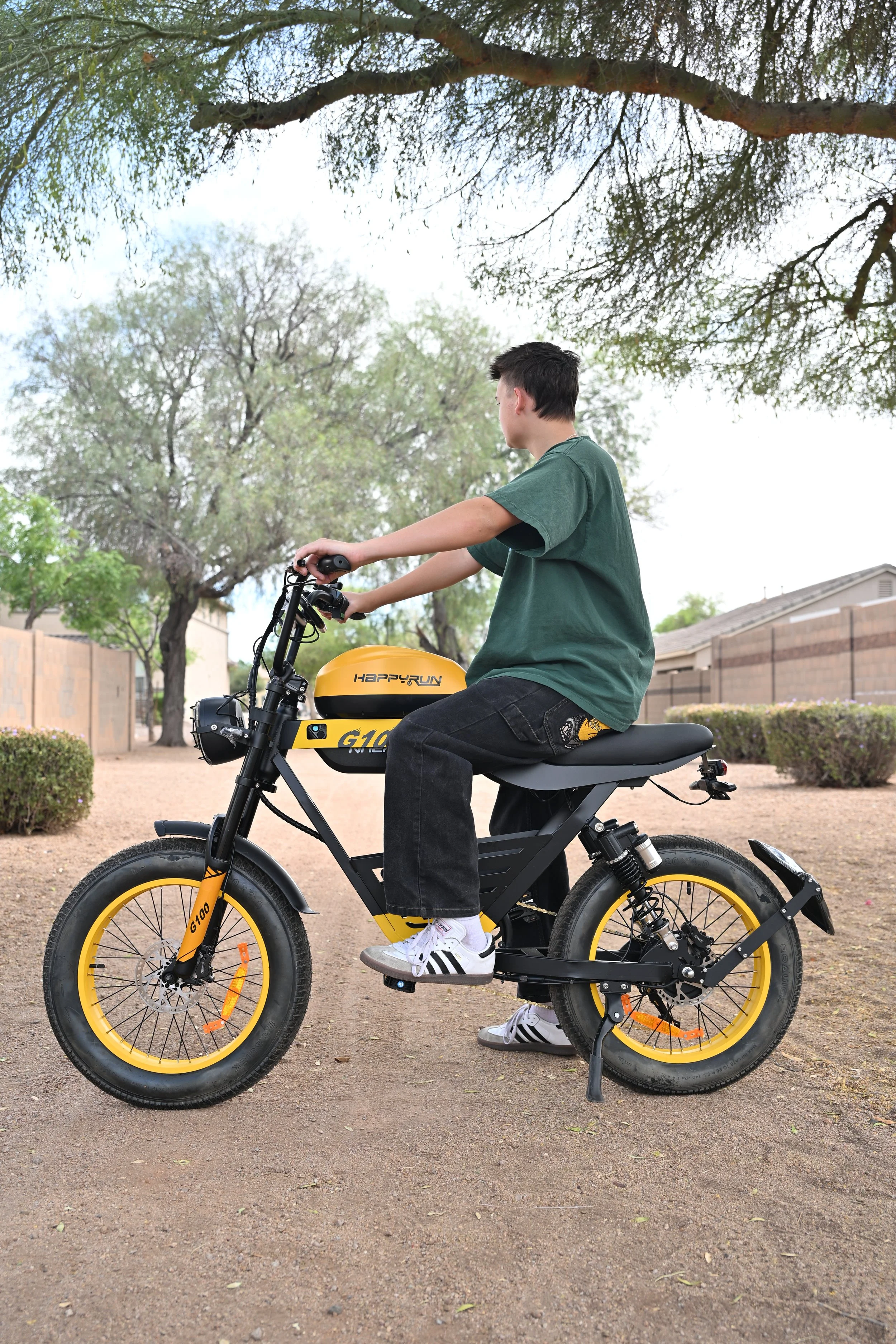 A young boy is sitting on a black and yellow electric bicycle in a park with trees and a residential fence in the background.