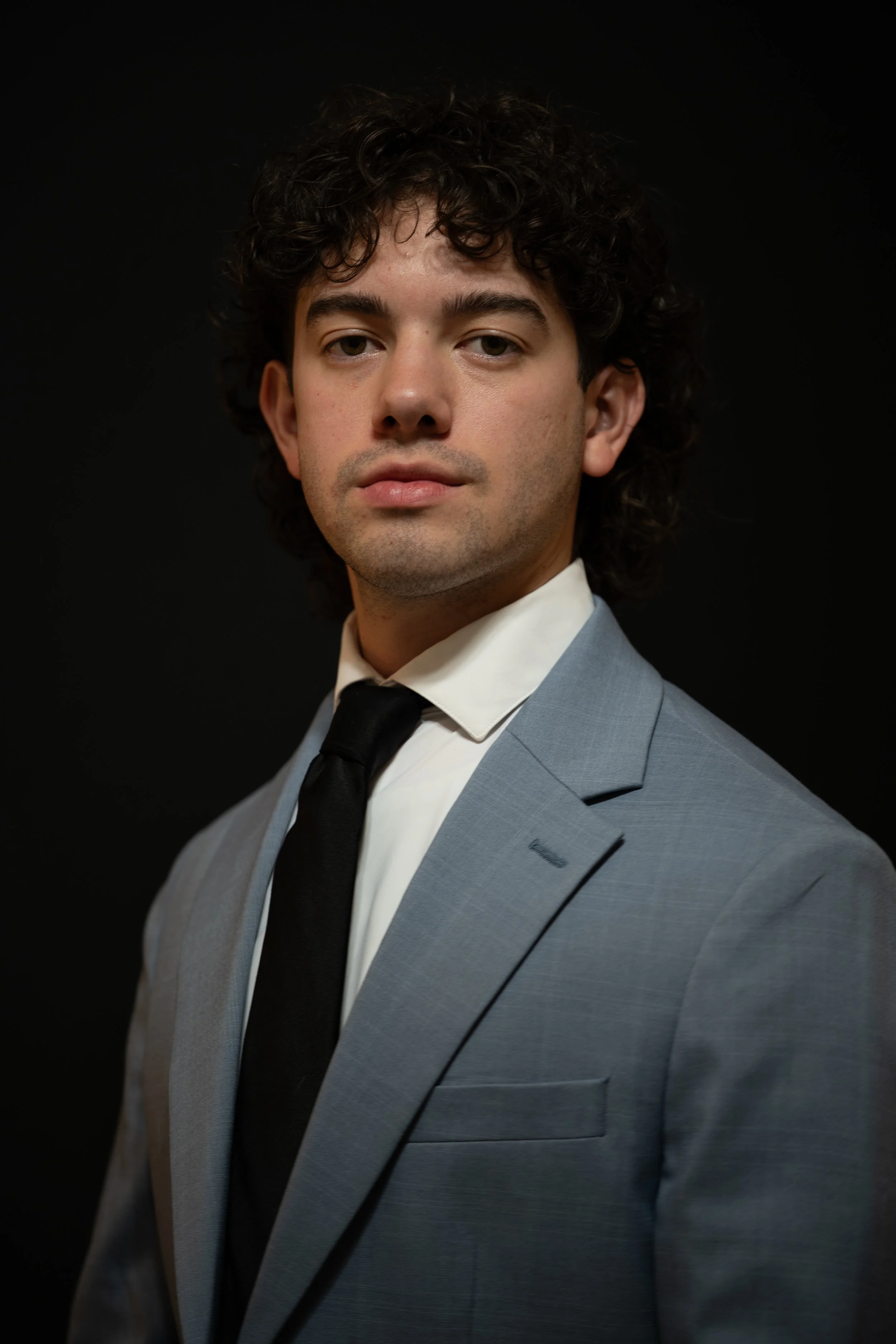 Portrait of a young man with curly dark hair, dressed in a light gray suit, white shirt, and black tie, against a black background.