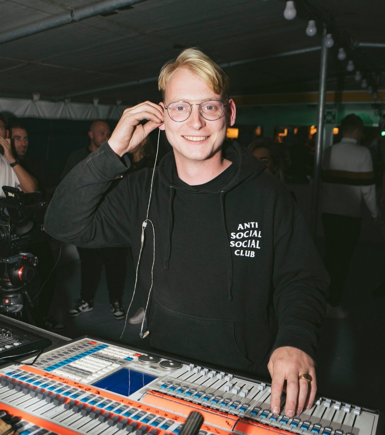 Person wearing glasses and a black "Anti Social Social Club" hoodie, standing at a sound mixing board, smiling and holding an earbud, with people in the background.