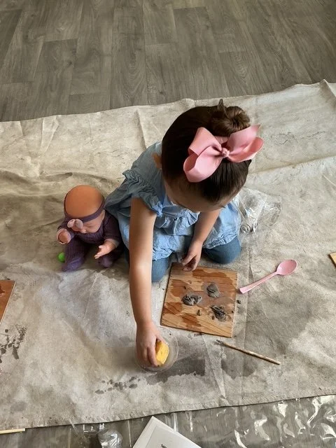 Una niña pequeña y una muñeca sentadas en una manta en el piso, con juguetes de arcilla y utensilios de modelado, jugando y creando arte.