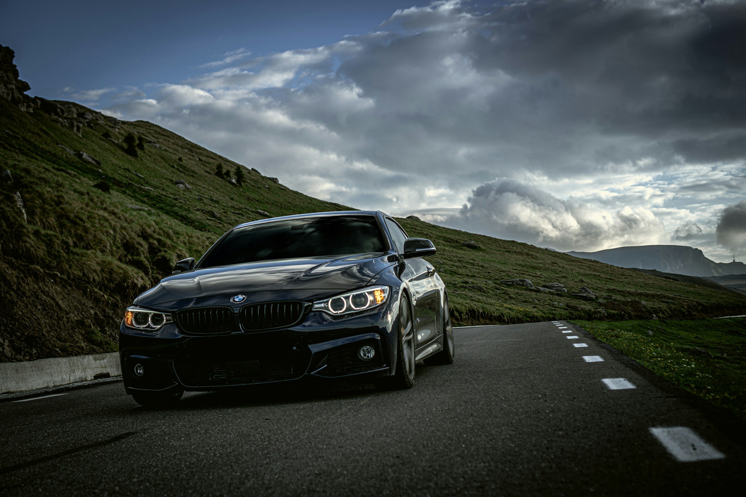 A black BMW car parked on the side of a mountain road with lush green hills and a dark cloudy sky in the background.