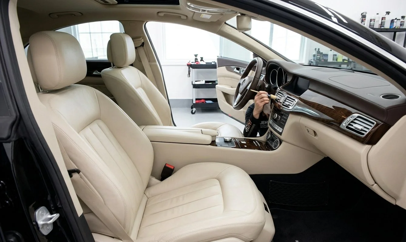 Interior view of a luxury car showing front seats, dashboard, and steering wheel. A person inside the car is holding a cleaning tool.