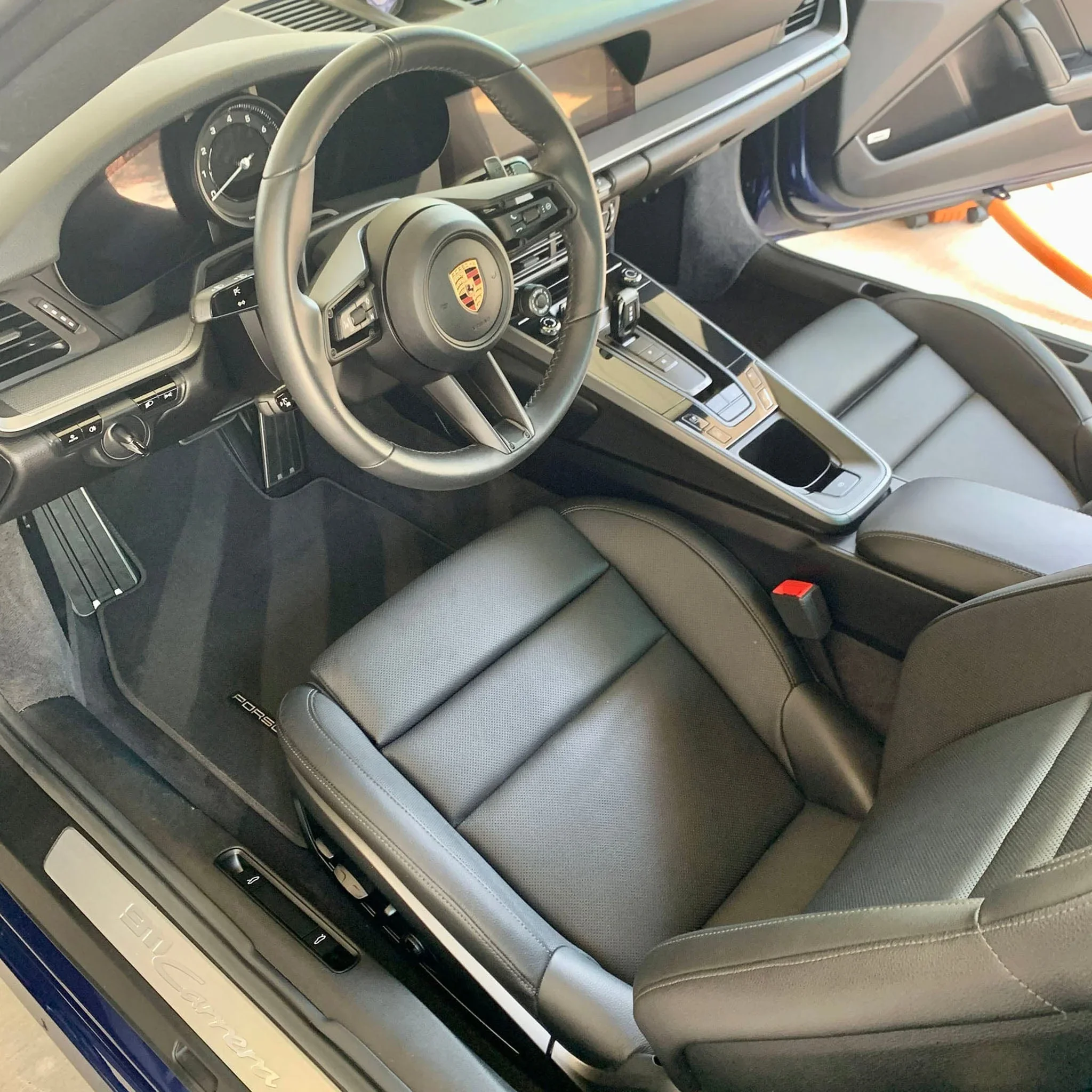 The interior of a Porsche vehicle, showing the steering wheel with the Porsche emblem, dashboard, gear shift, and black leather seats.