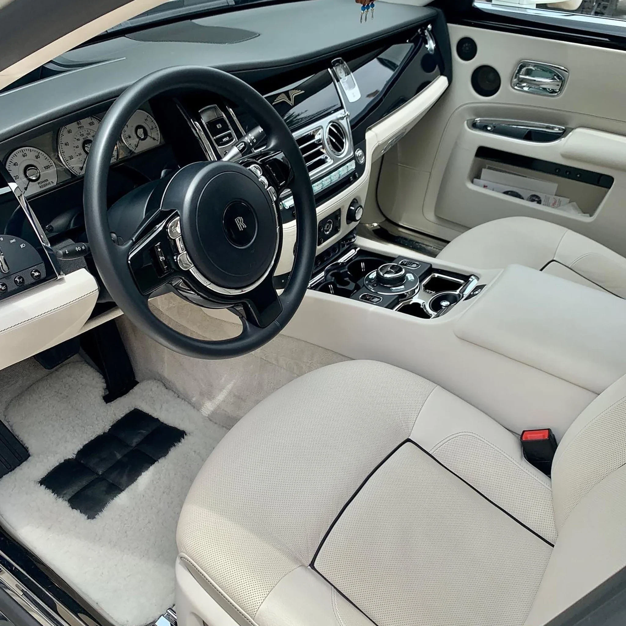The interior of a luxury car showing a beige leather seat, a black steering wheel with the Rolls-Royce emblem, a dashboard with various gauges, and a central console with controls, all in a high-end vehicle.
