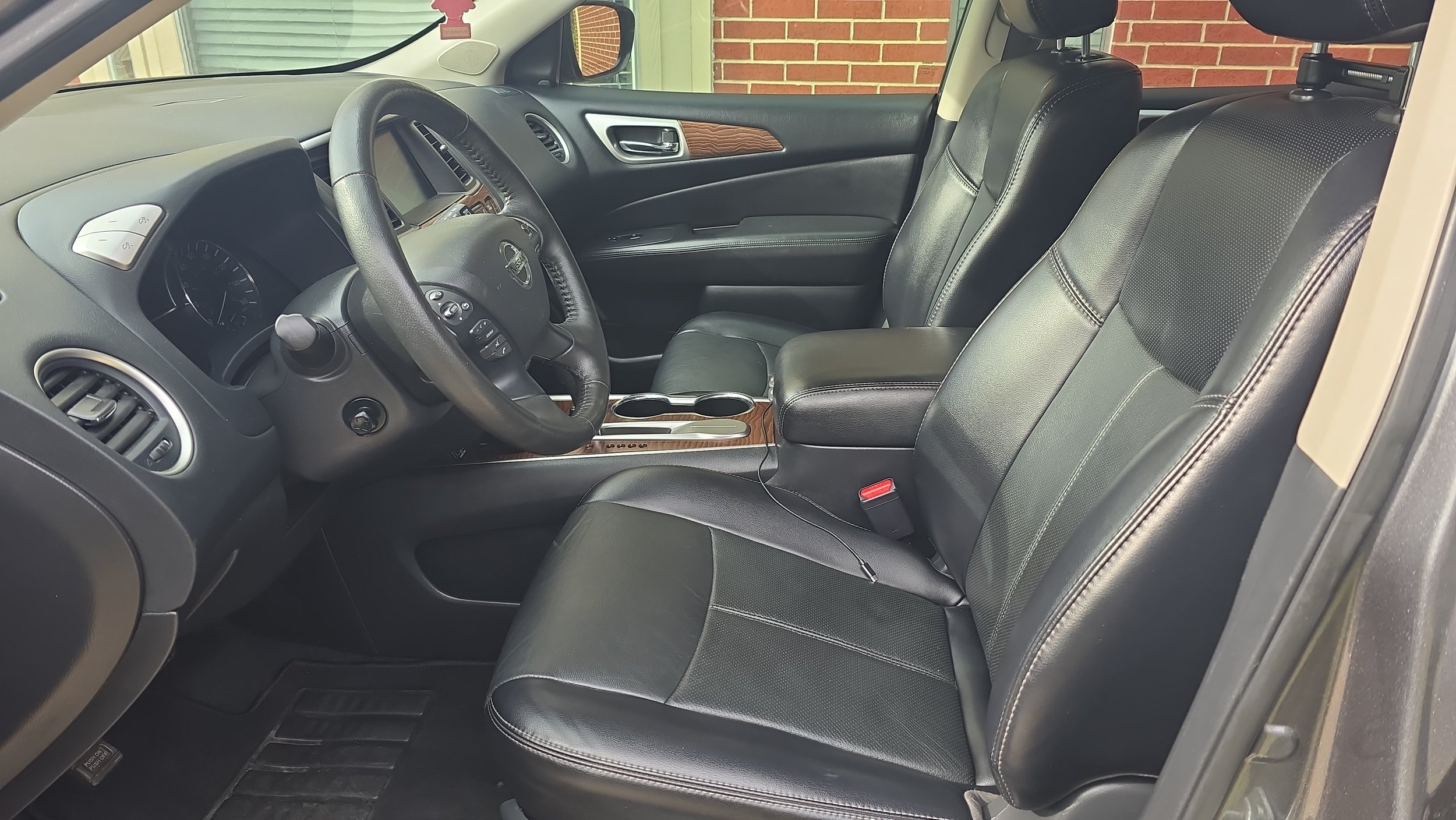The interior of a car showing the front black leather seats, steering wheel, dashboard, and center console with wood grain accents, and part of the brick wall outside visible through the window.