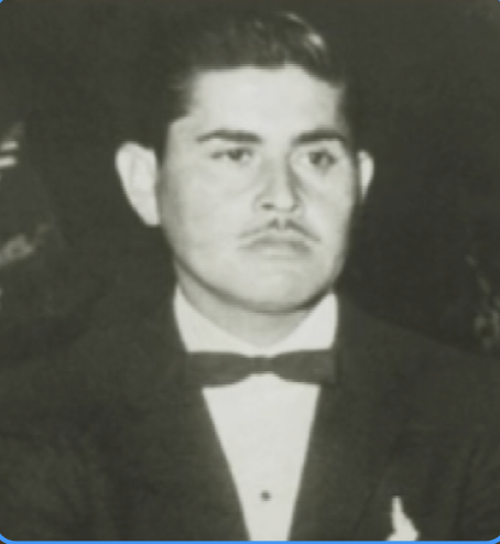 Retrato en blanco y negro de un hombre joven con peinado clásico, vestimenta formal con saco y pajarita, en un entorno que parece ser una celebración o evento formal.