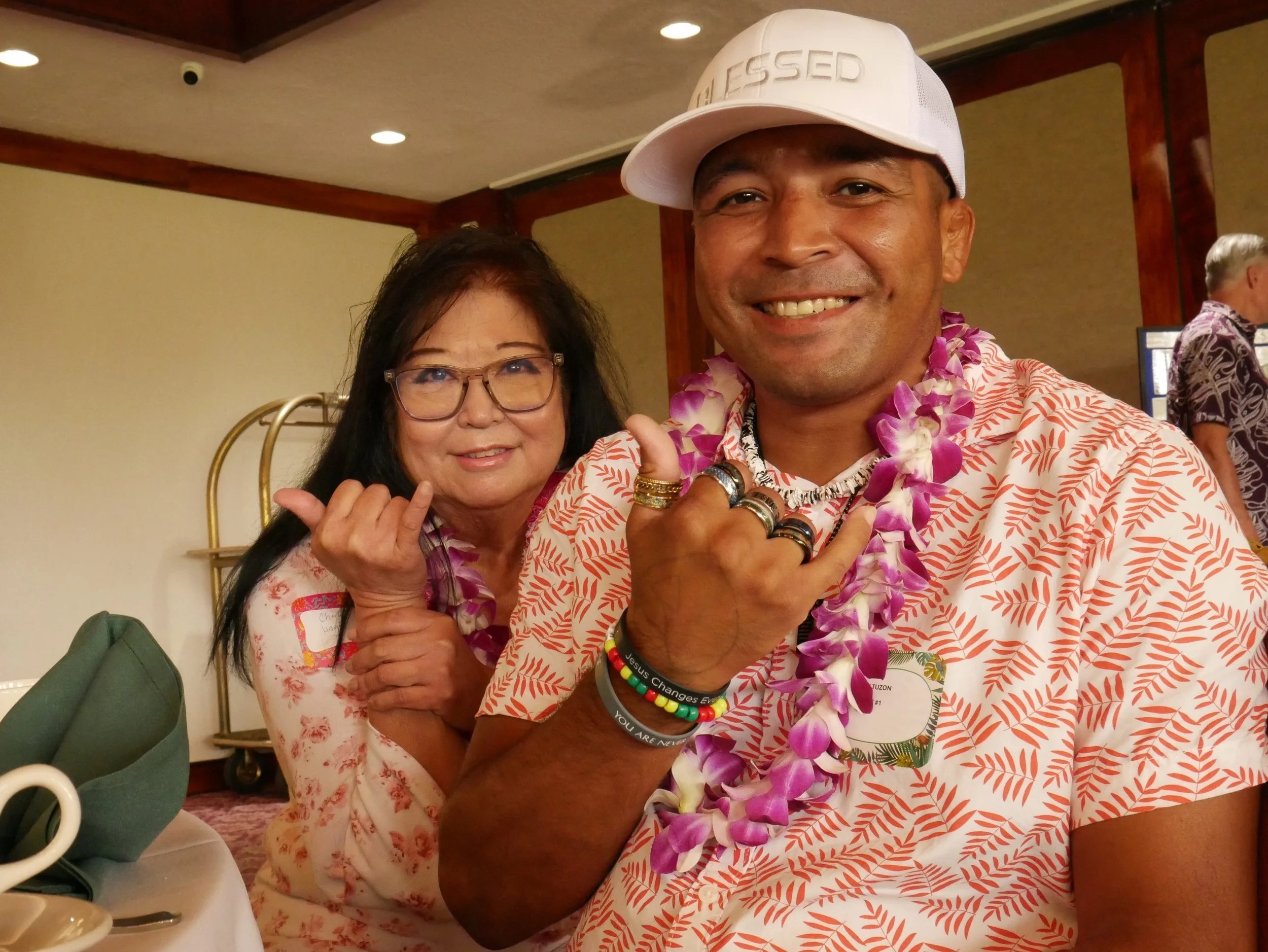 Two people smiling at a tropical-themed event, both making the 'shaka' hand gesture. The man is wearing a white cap, lei, and tropical shirt, and the woman is wearing glasses and a floral top.