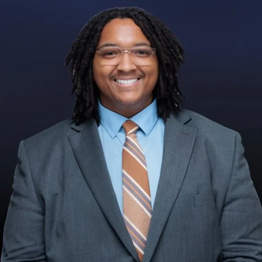 Portrait of a man in a gray suit, light blue shirt, and striped tie, smiling against a dark background.