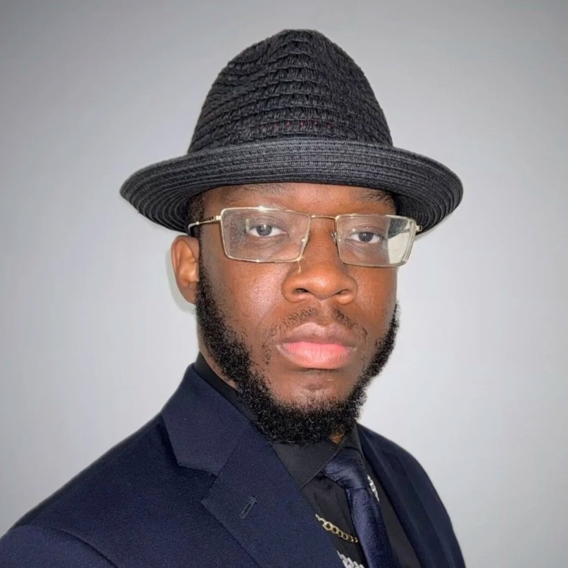 A man in a suit with a hat and glasses, posing against a plain background.
