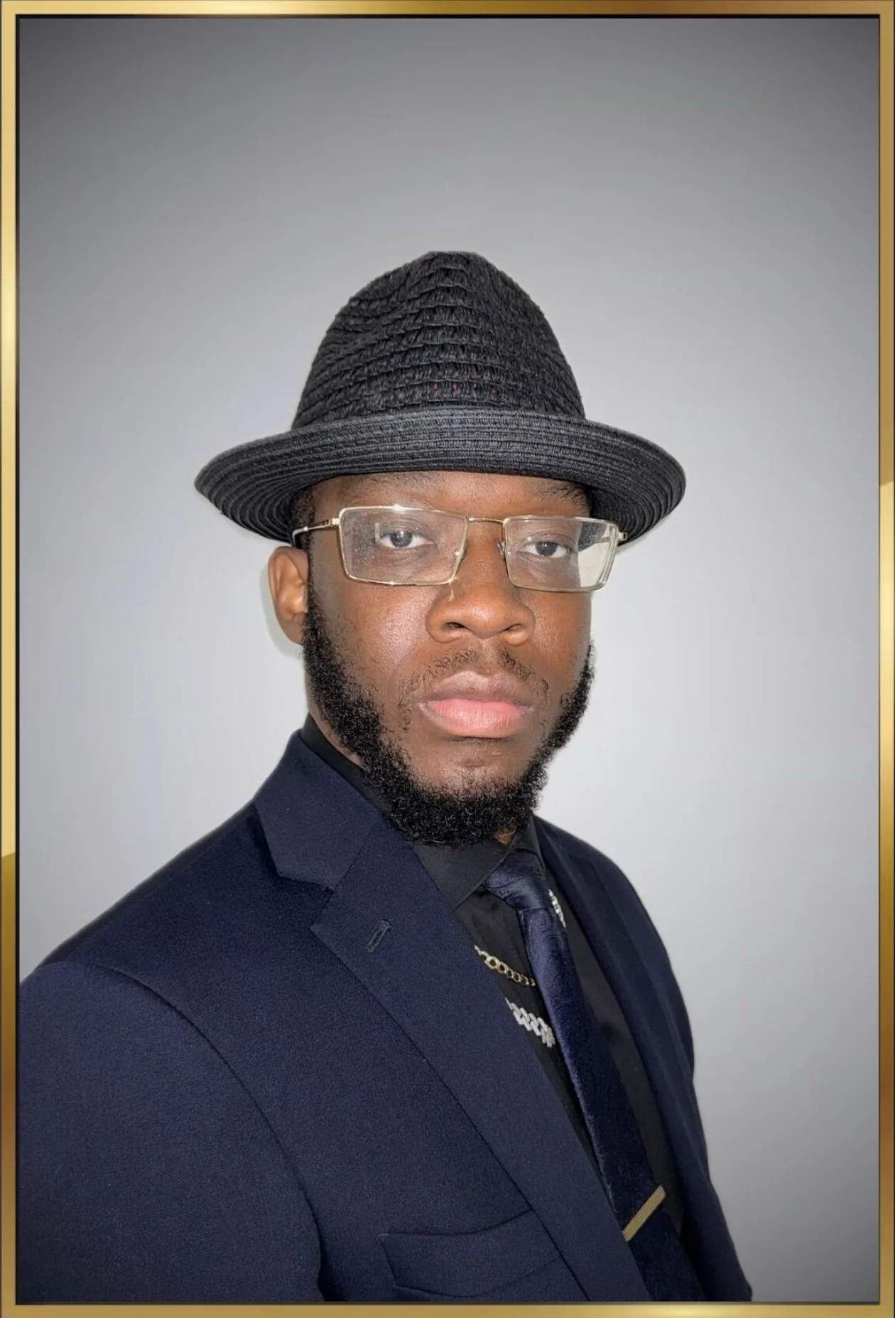 Man wearing a black hat, glasses, a navy suit, and a tie, posing against a plain white background.