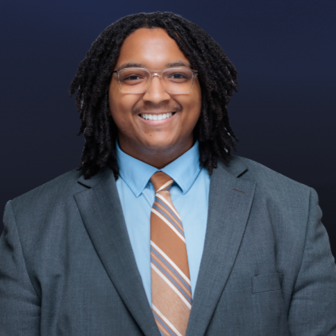 Smiling man wearing glasses, a gray suit, light blue shirt, and striped tie, standing against a dark background with a gold border.