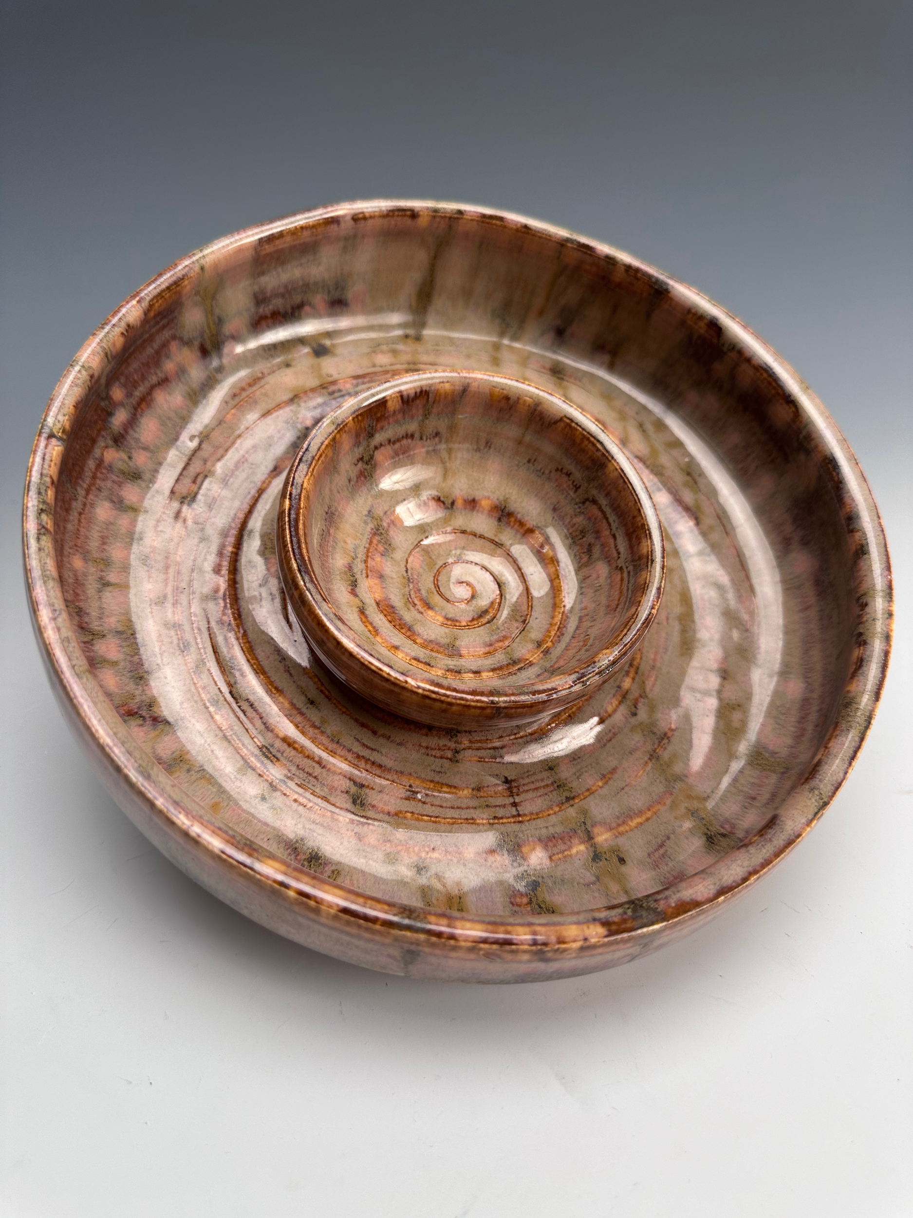 A spiral-shaped ceramic dish with earthy tones and a glossy finish, photographed from above on a white background.
