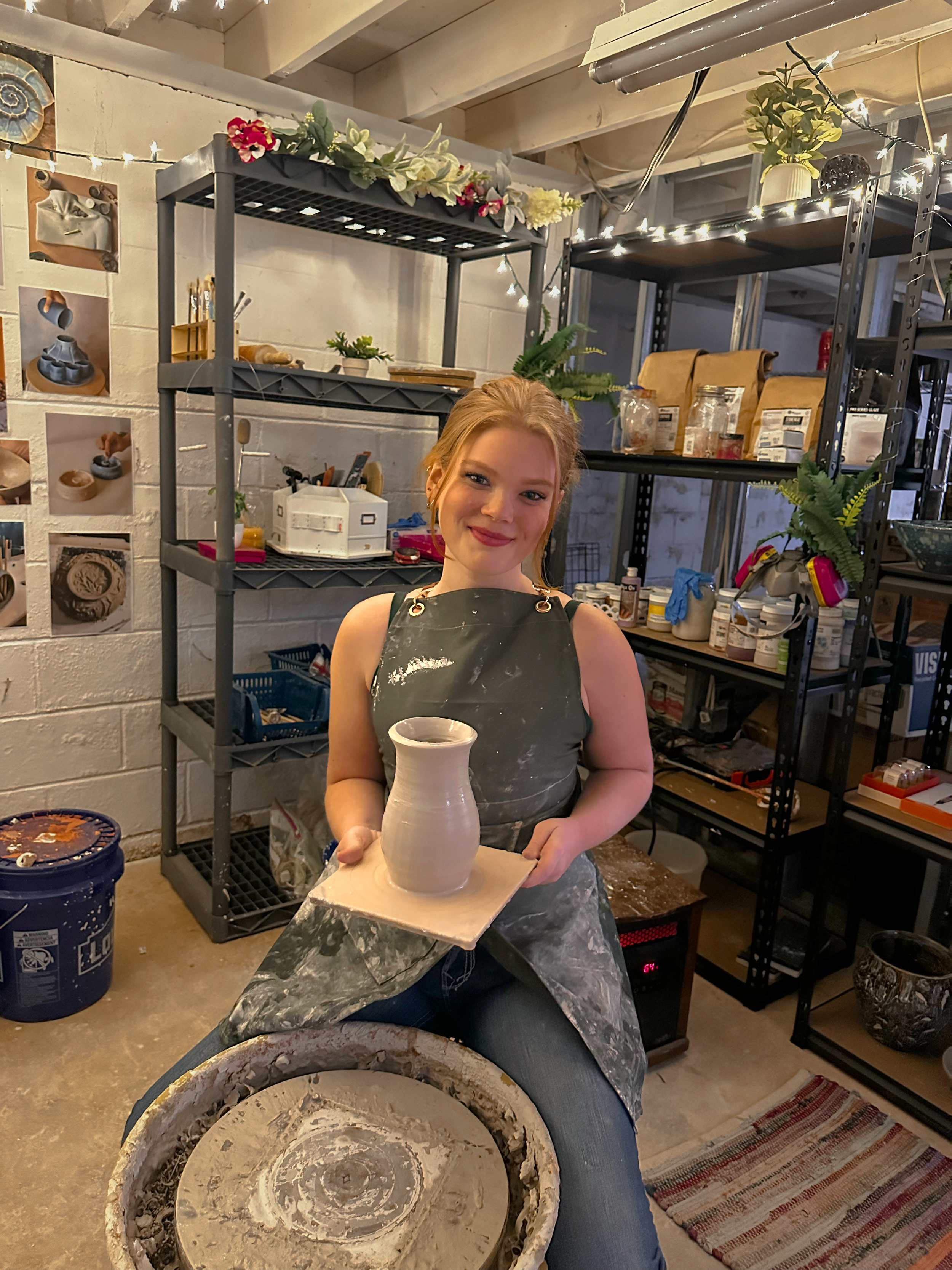 A young woman with red hair smiling and holding a ceramic vase in a pottery studio. She is wearing a black apron and sitting in front of a potter's wheel. The studio has shelves with bags of clay, jars, plants, and decorative string lights.