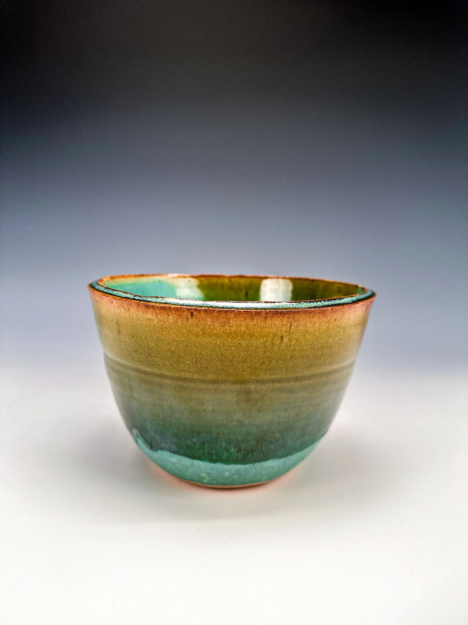 A ceramic bowl with a multicolored glaze, featuring green, brown, and turquoise hues, against a gradient background that transitions from dark at the top to light at the bottom.