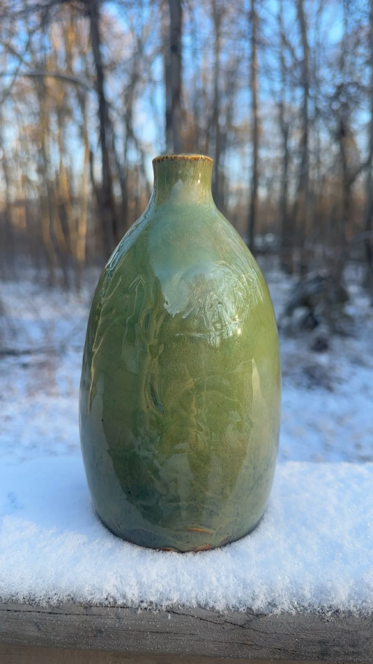 A green glazed ceramic vase placed on a snow-covered surface outdoors, with a background of leafless trees and a blue sky.