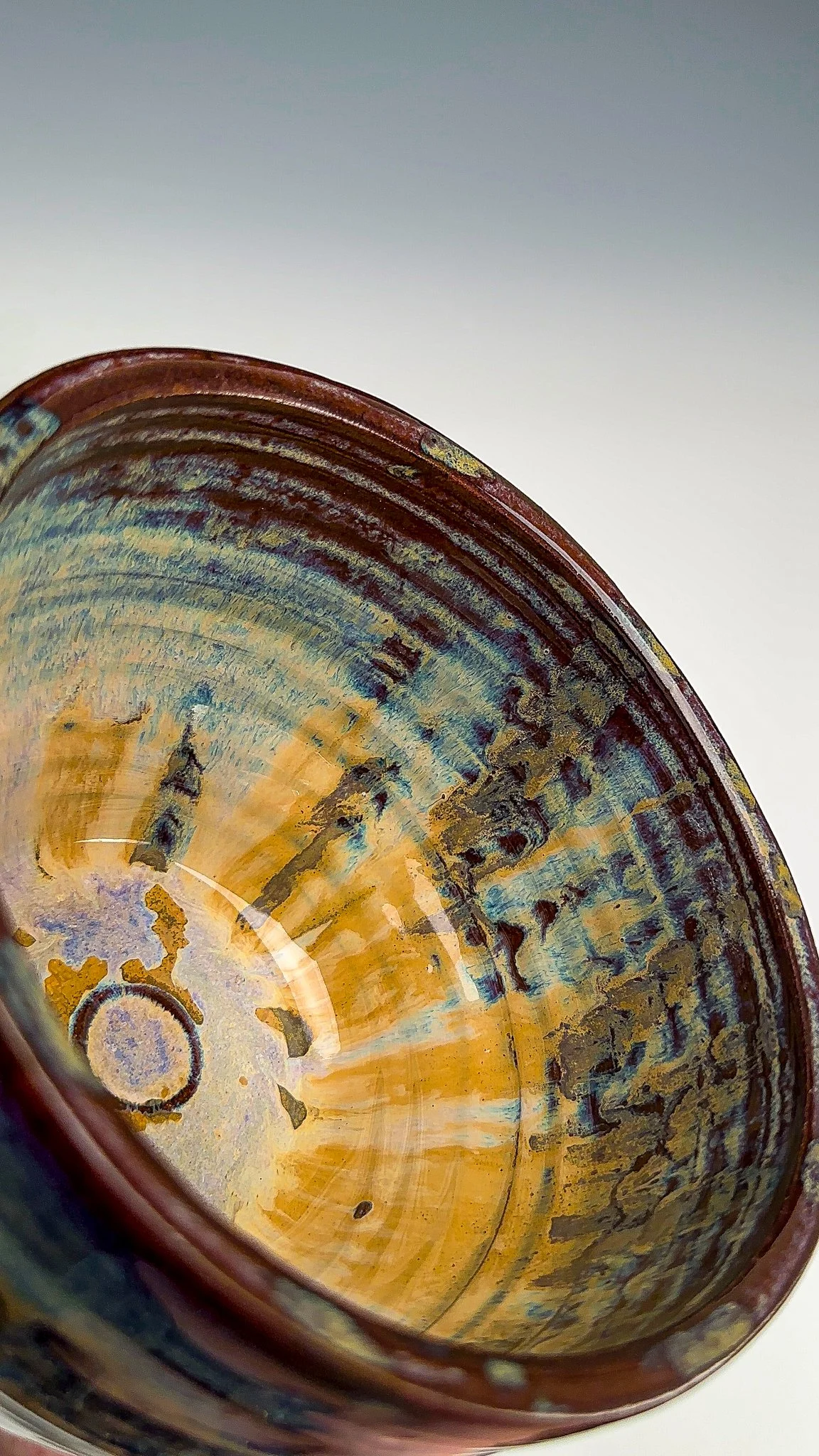 Close-up view of a ceramic cup with a glazed interior featuring abstract patterns in shades of blue, green, yellow, and orange, with a plain gray background.