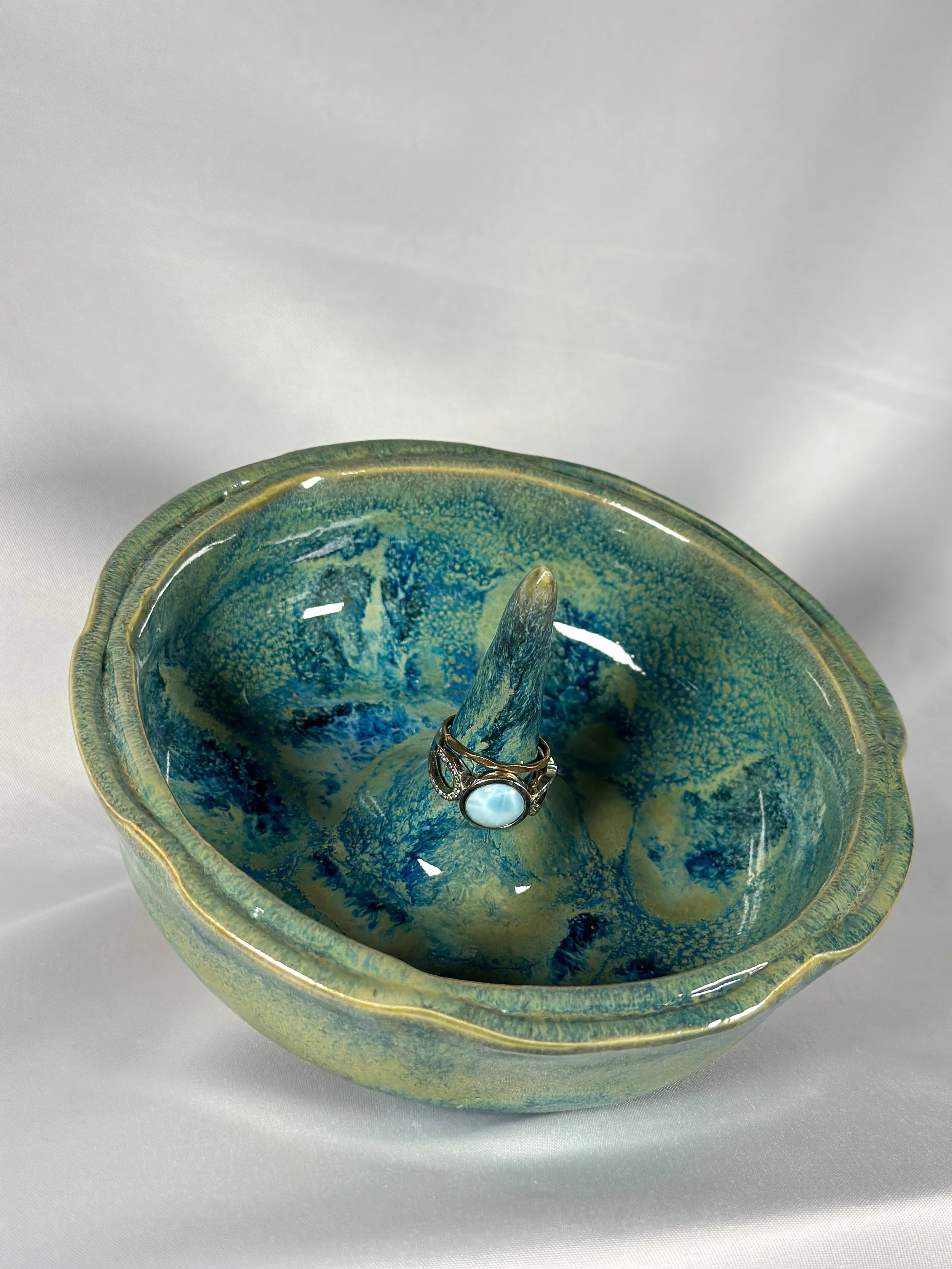 A ceramic bowl with blue and green glaze, holding a ring with a large white pearl and intricate silver detailing.