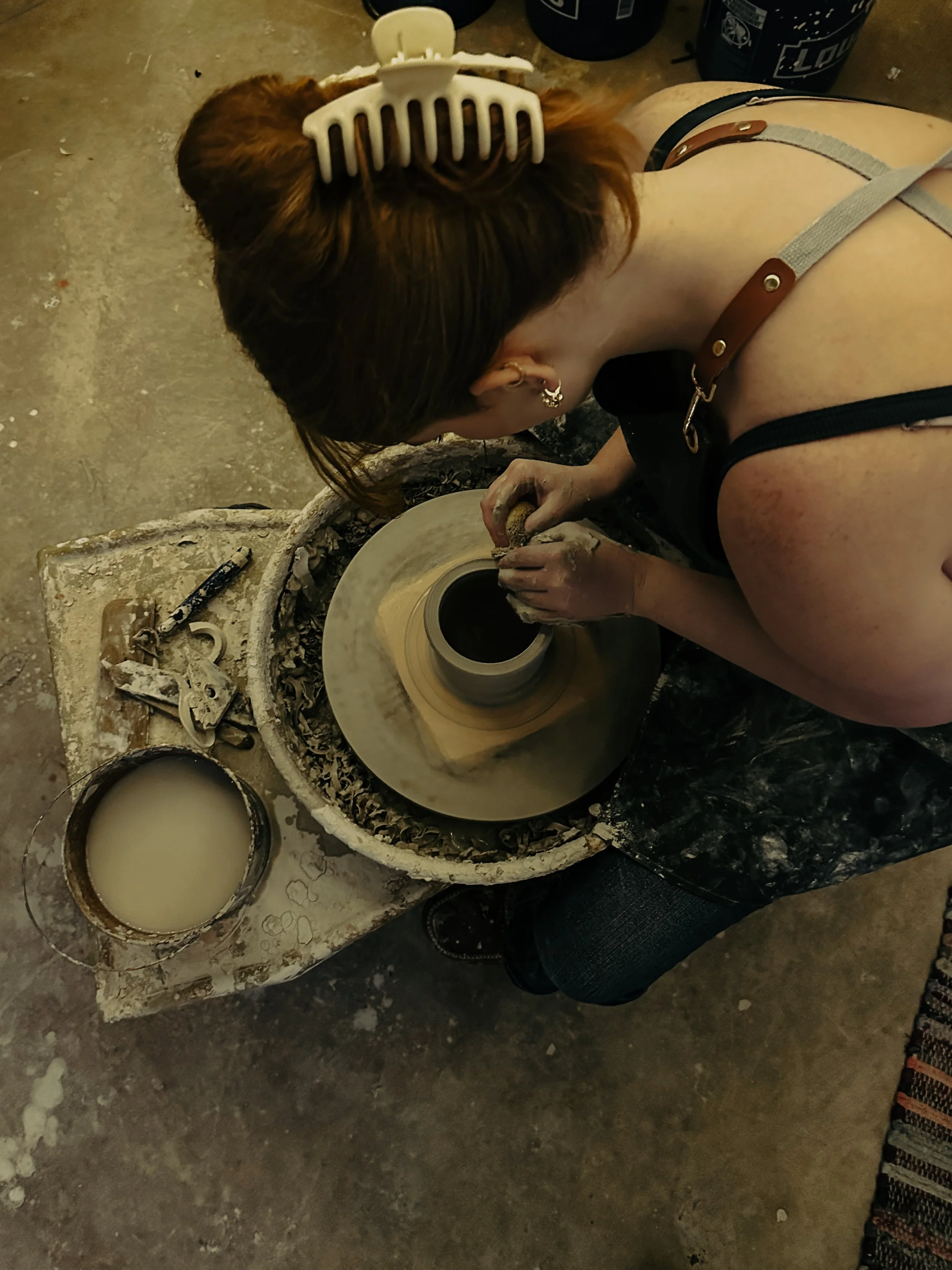 A person with red hair, wearing a hair clip, is shaping clay on a pottery wheel in a studio. The person's hands are covered in clay, and there are tools and a bucket of water on a nearby work table.