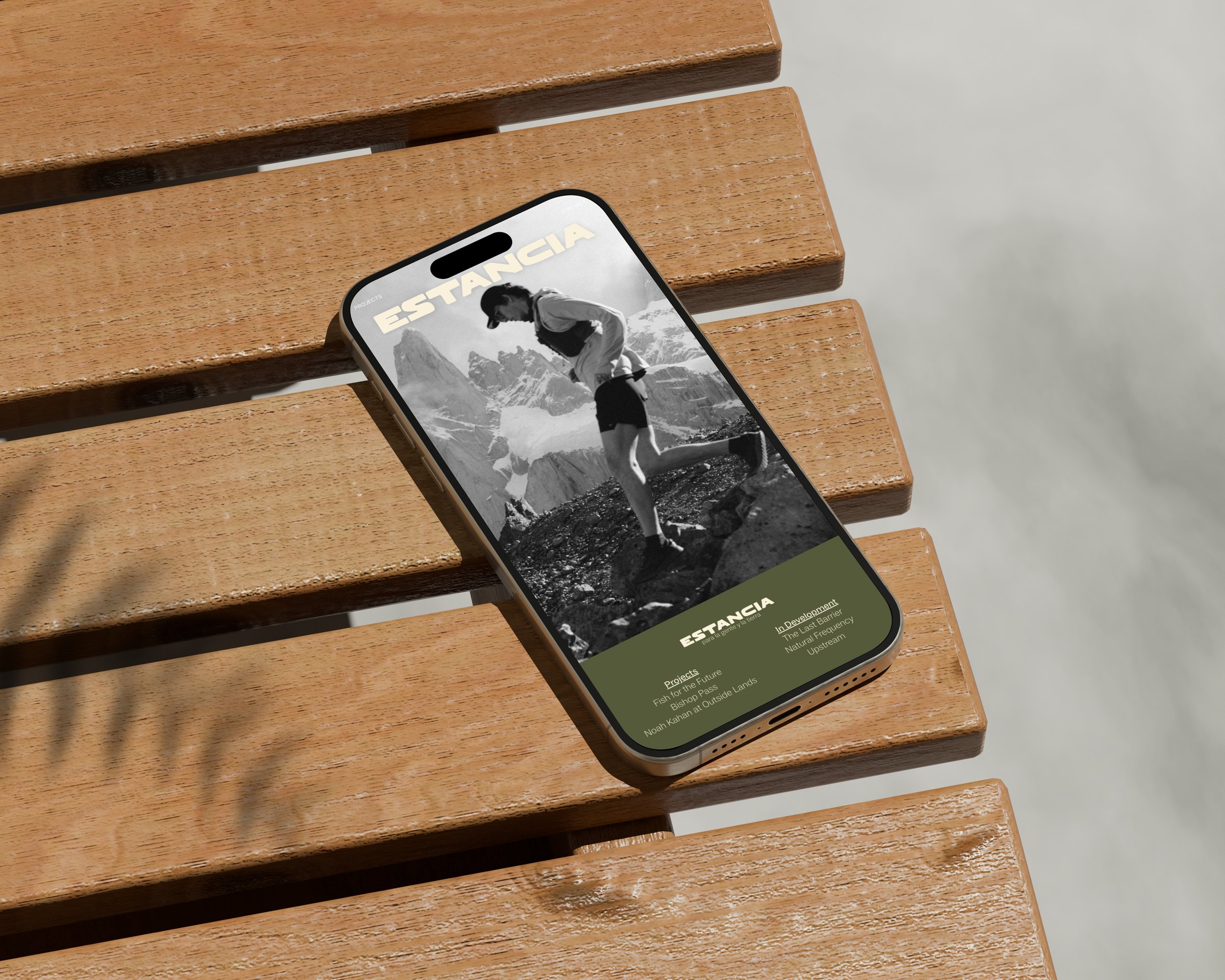 A smartphone resting on wooden benches displays a black and white photo of a hiker in front of mountainous terrain and text about various projects.