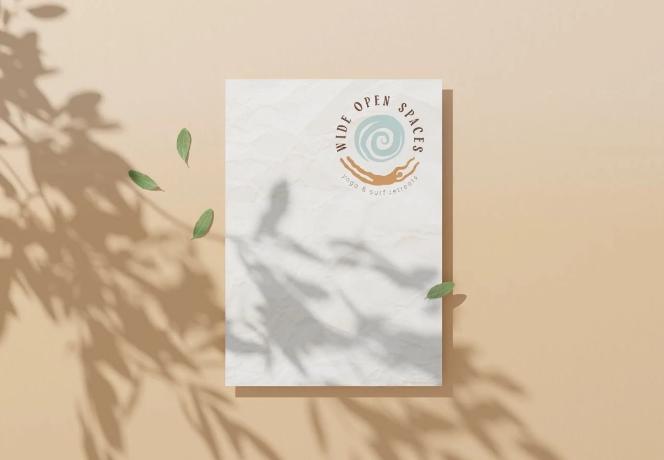 A printed sign that reads "WIDE OPEN SPACES Yoga & Surf Retreats" with a circular logo featuring a spiral, a hand, and a wave. The sign is on a beige wall with shadows of leaves cast on it.