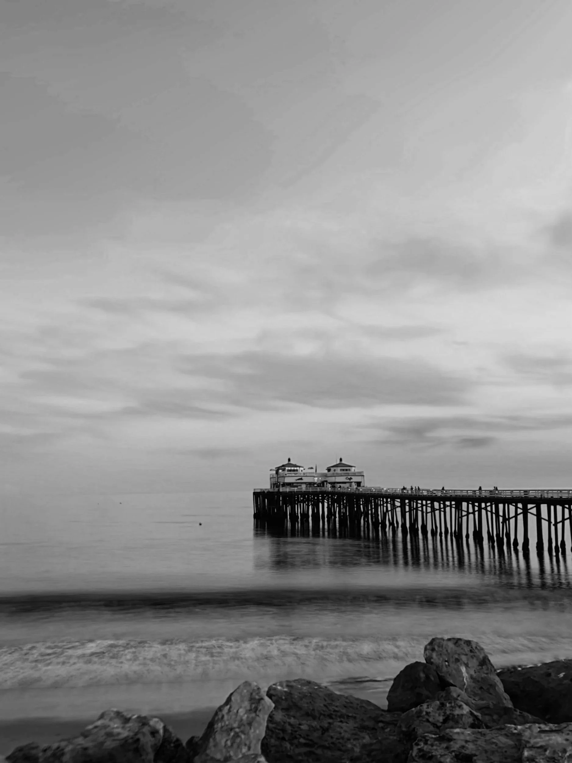 A black and white photo of a pier extending into calm water, with a rocky beach in the foreground and a cloudy sky.