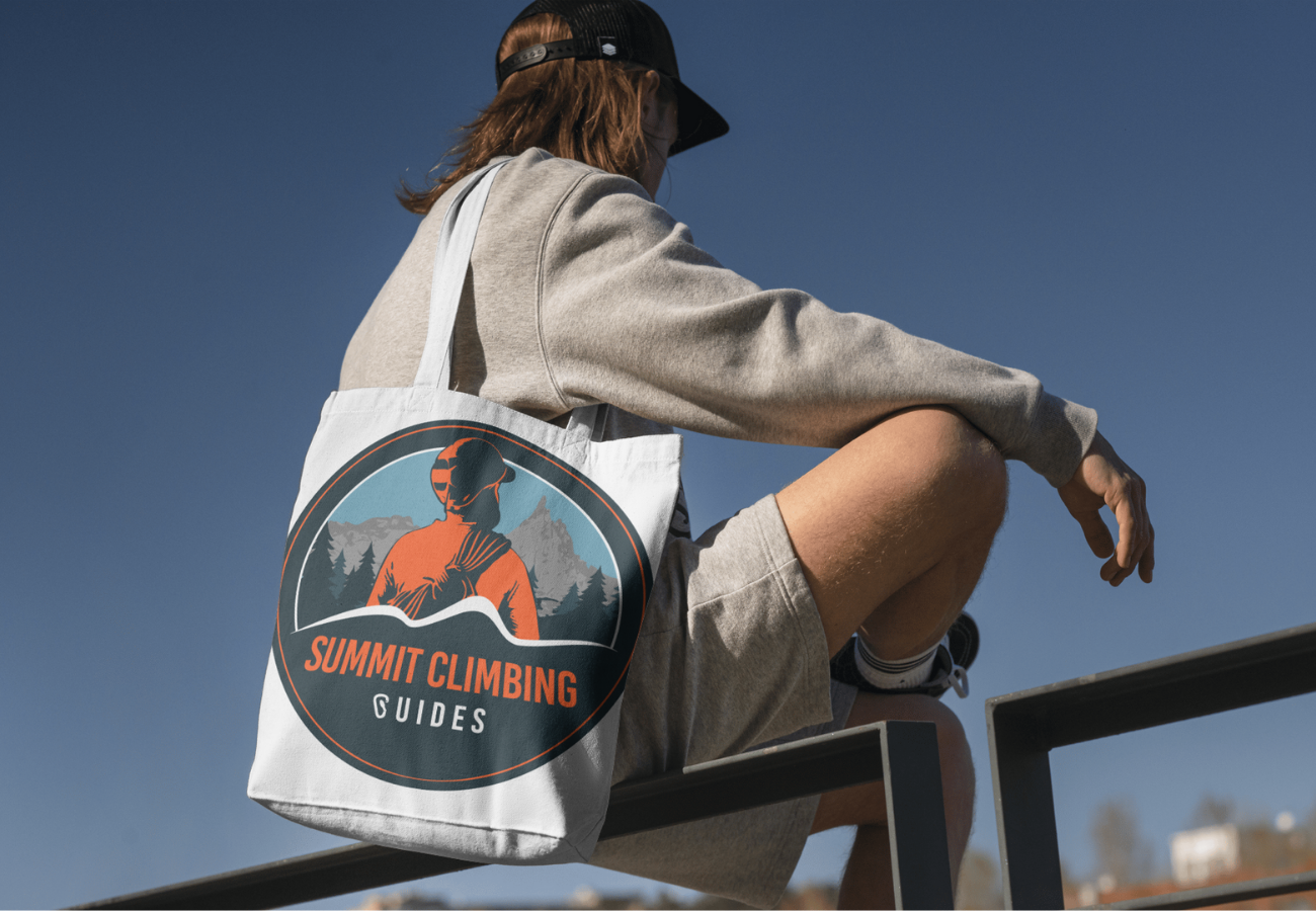 Person sitting on a ledge wearing a cap, grey sweatshirt, and shorts, carrying a tote bag with a mountain climbing logo, under a clear blue sky.