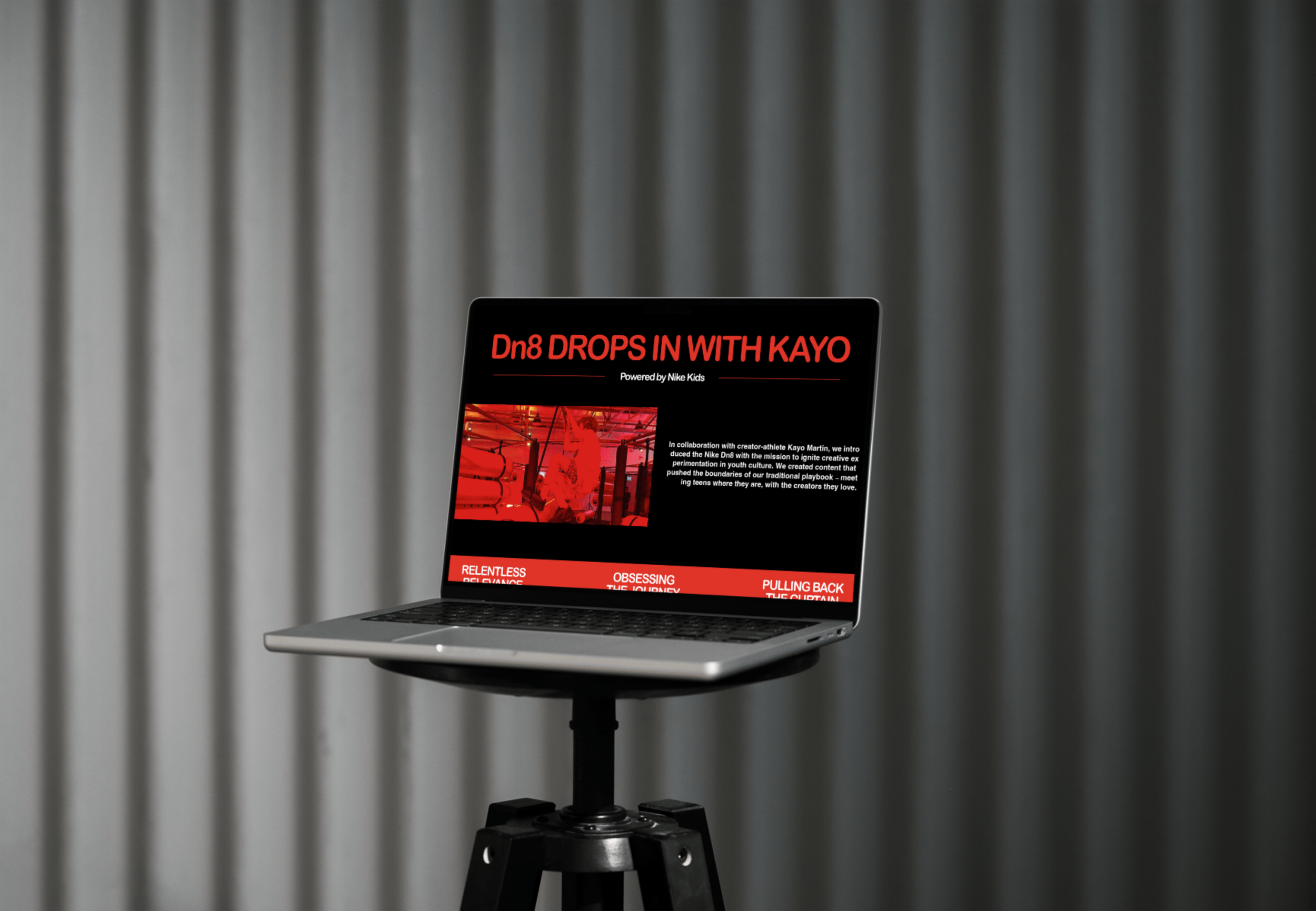 A laptop on a stand displays a website titled "Dn8 DROPS IN WITH KAYO" with red and black design elements, text, and a small image, set against a gray, curtain-like background.