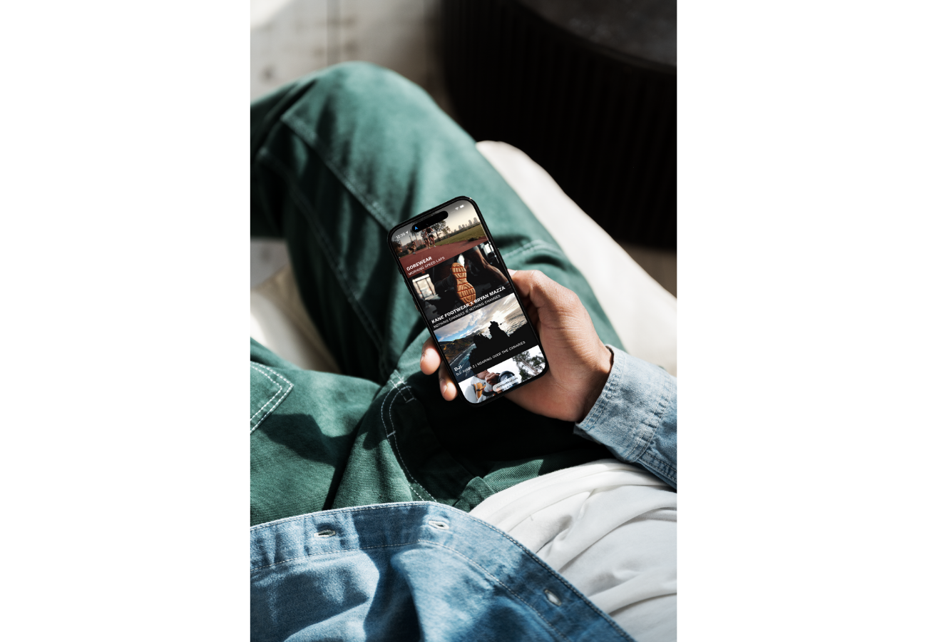 Person sitting with legs crossed, wearing green pants and a denim jacket, holding a smartphone displaying a fashion website or app.